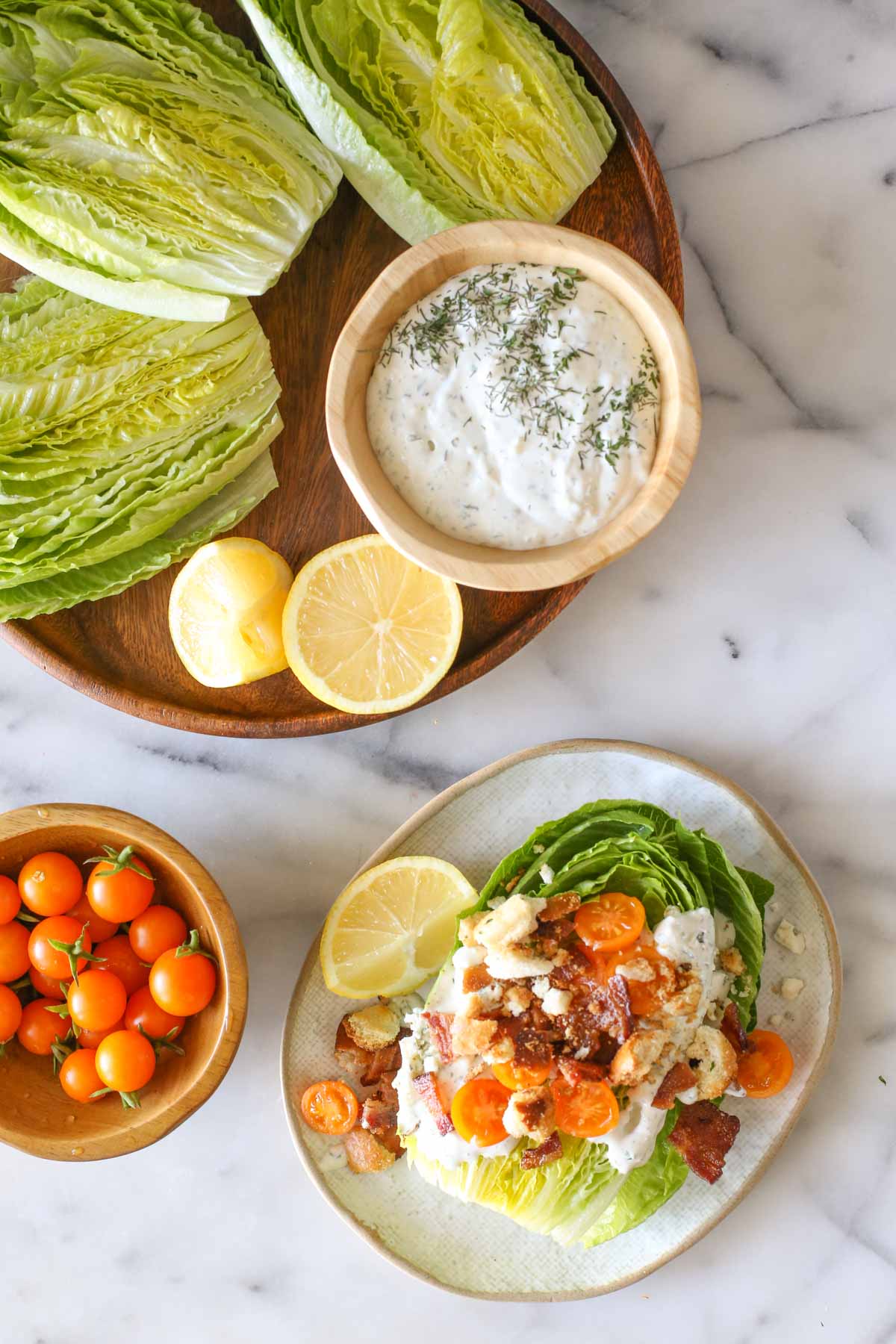 Overhead shot of a wood platter with three romaine lettuce halves, lemon wedges and a wood bowl of the blue cheese dressing on it, sitting next to a wood bowl of Sungold tomatoes, and a Wedge Style BLT Salad on a plate, all on a marble background.  