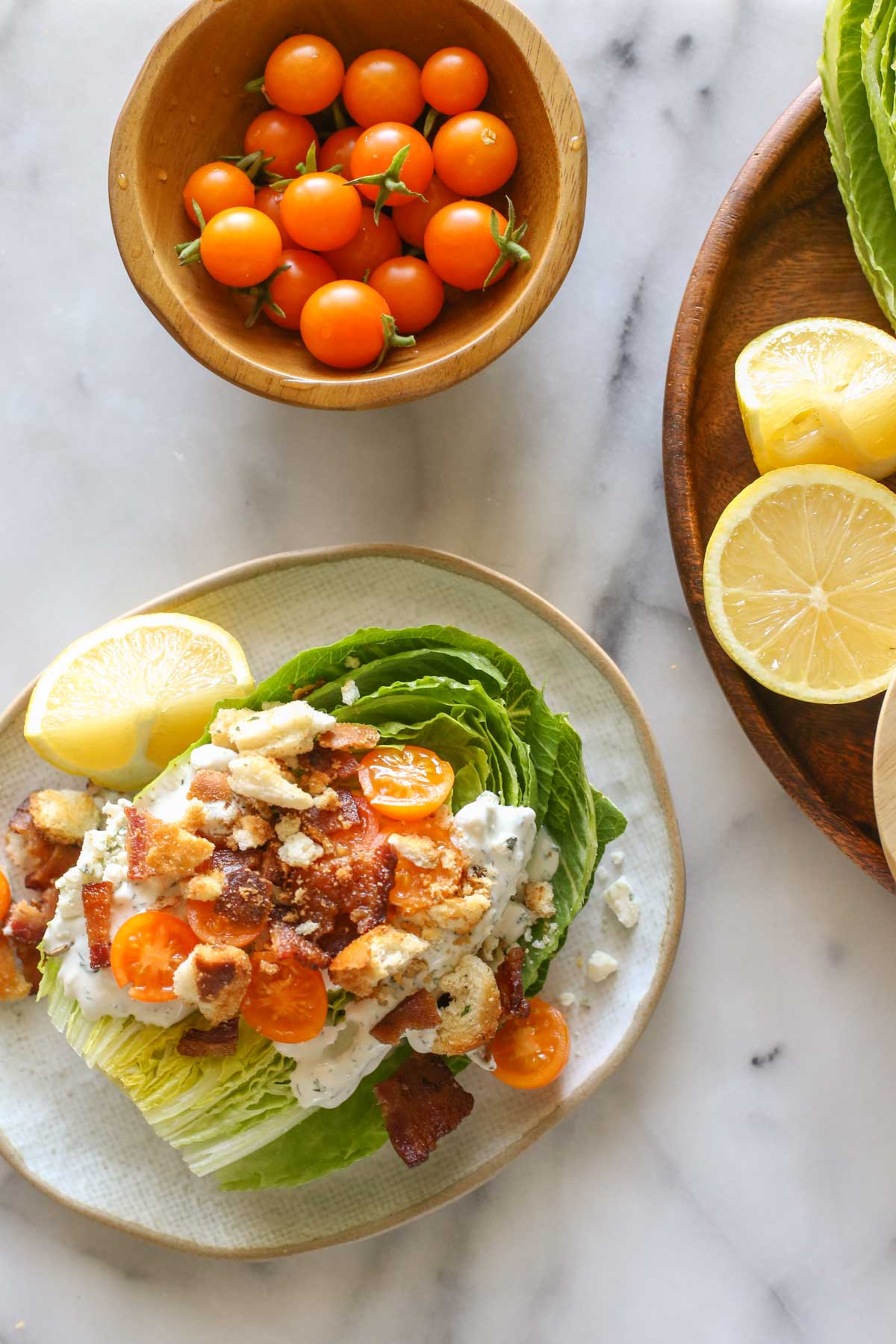 Overhead shot of a Wedge Style BLT Salad on a plate with a lemon wedge, next to a wood bowl of Sungold tomatoes and a wood platter with lemon wedges, all on a marble background. 