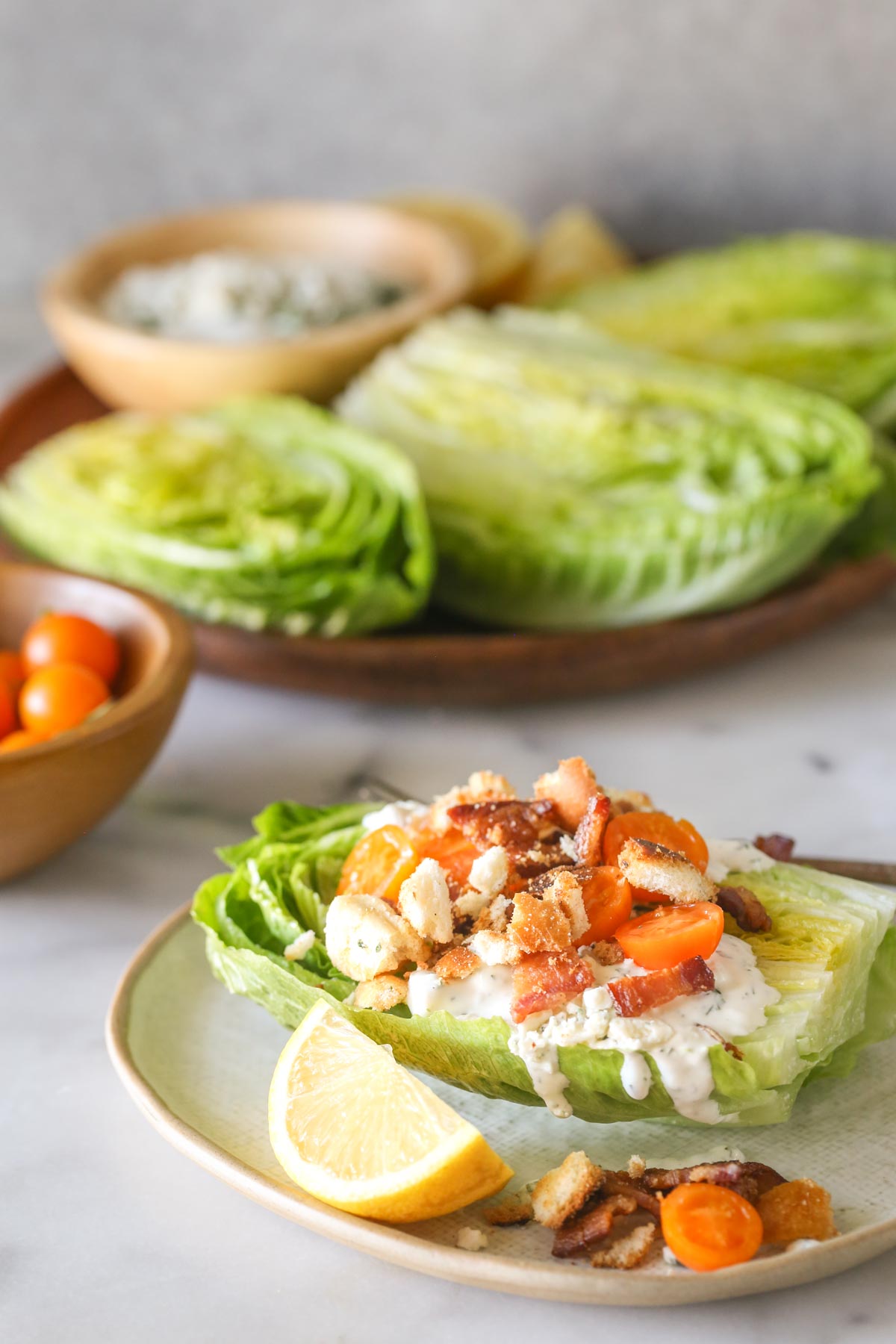 A Wedge Style BLT Salad on a plate with a lemon wedge, with a wood bowl of Sungold tomatoes and a wood platter with the romaine lettuce halves, a bowl of blue cheese dressing and lemon wedges on it in the background. 