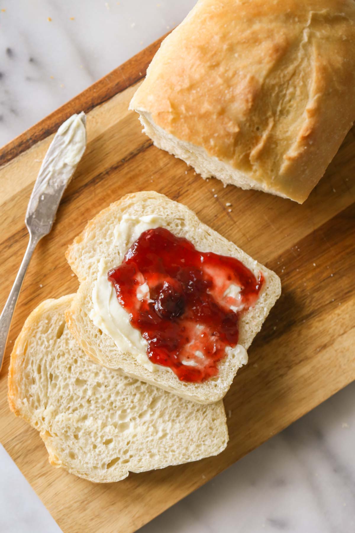Overhead shot of a Sourdough Sandwich Loaf sliced on a cutting board with a butter knife next to the bread, and one of the slices of bread has butter and jelly on it.  