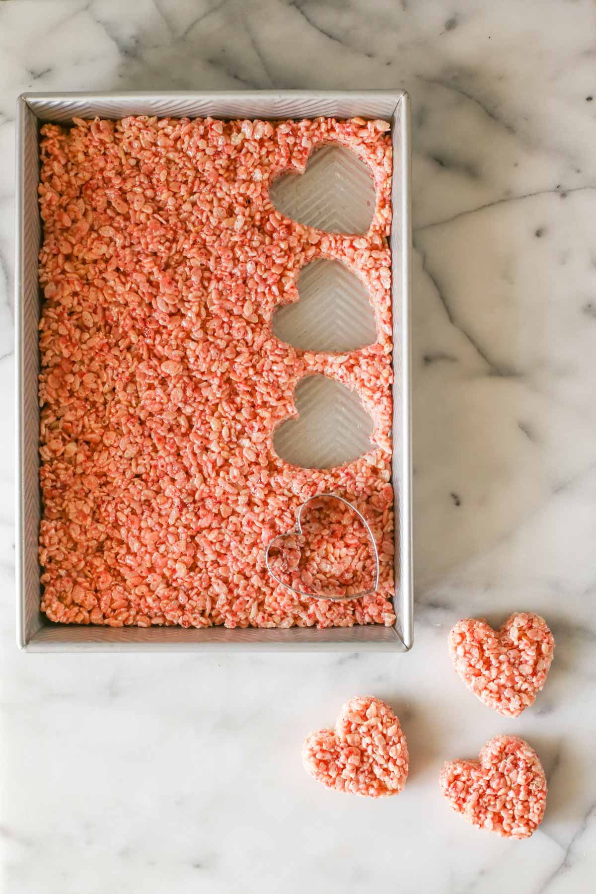 Overhead shot of a pan of Strawberry Rice Krispie Treats with a heart shaped cutter in the pan with three hearts already cut out and laying next to the pan, all on a marble background. 