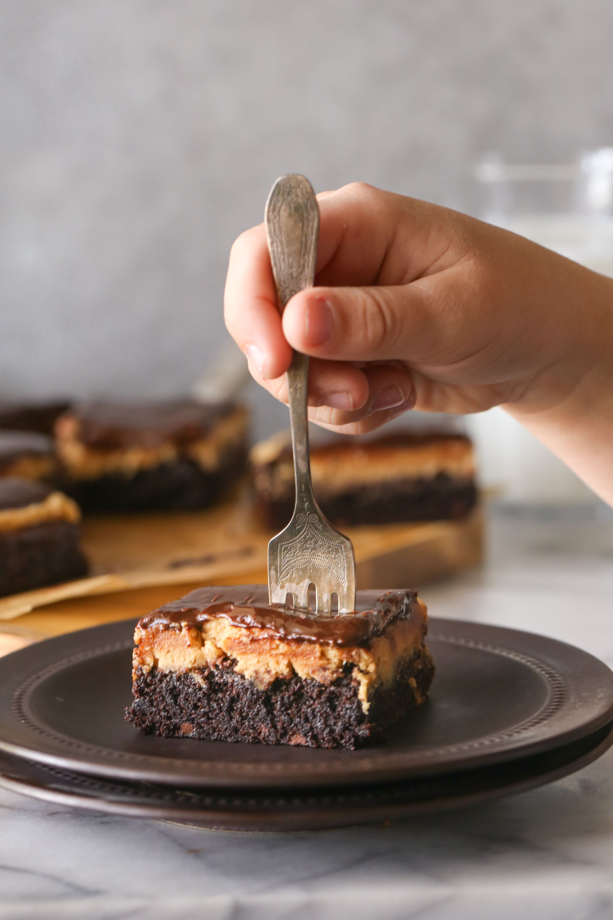 A Chocolate Peanut Butter Brownie on a plate with a hand holding a fork stuck in the middle of the brownie, with more brownies in the background. 