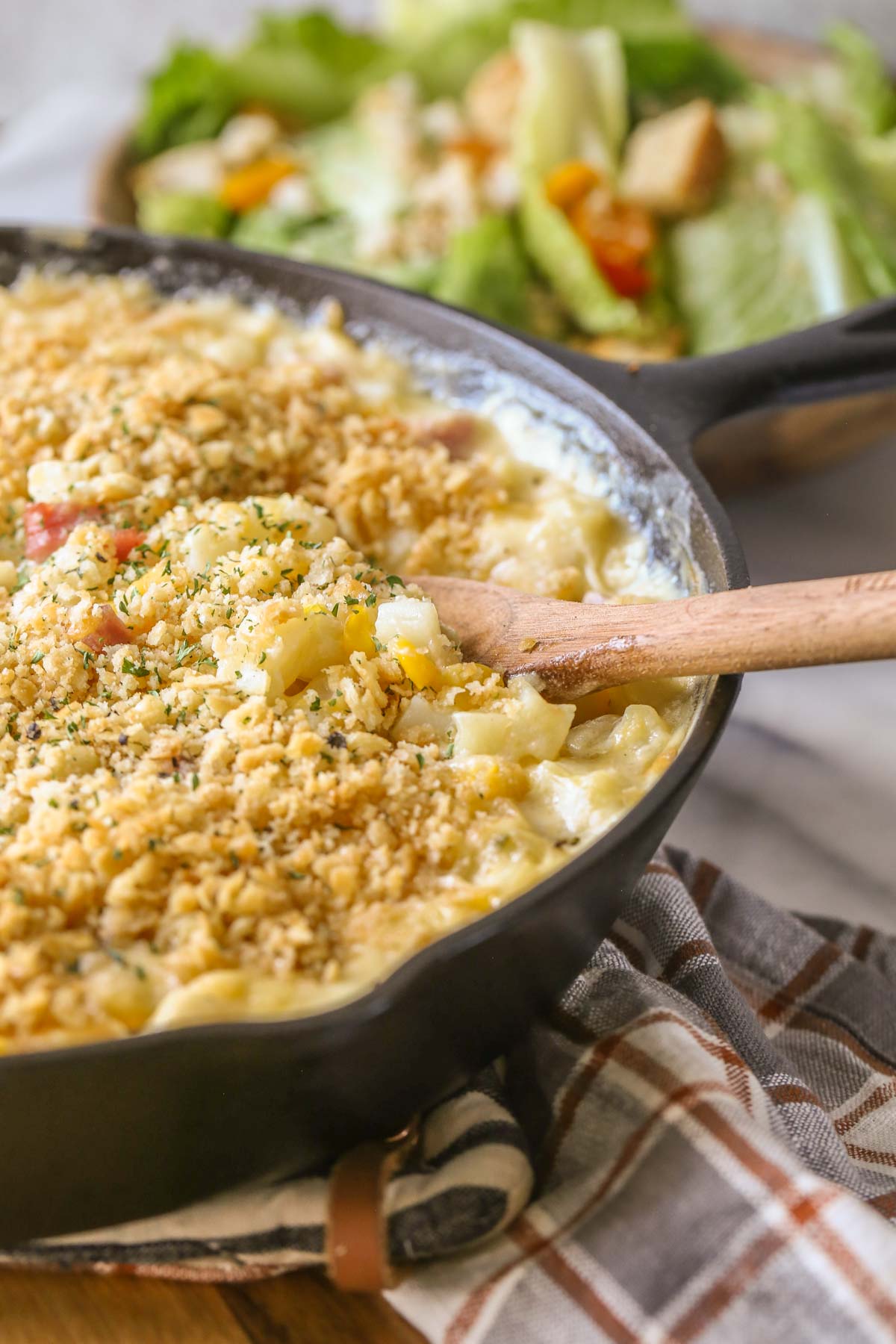 Close up shot of Cheesy Ham and Potato Skillet in a cast iron skillet with a wooden serving spoon in it, and a bowl of salad in the background. 