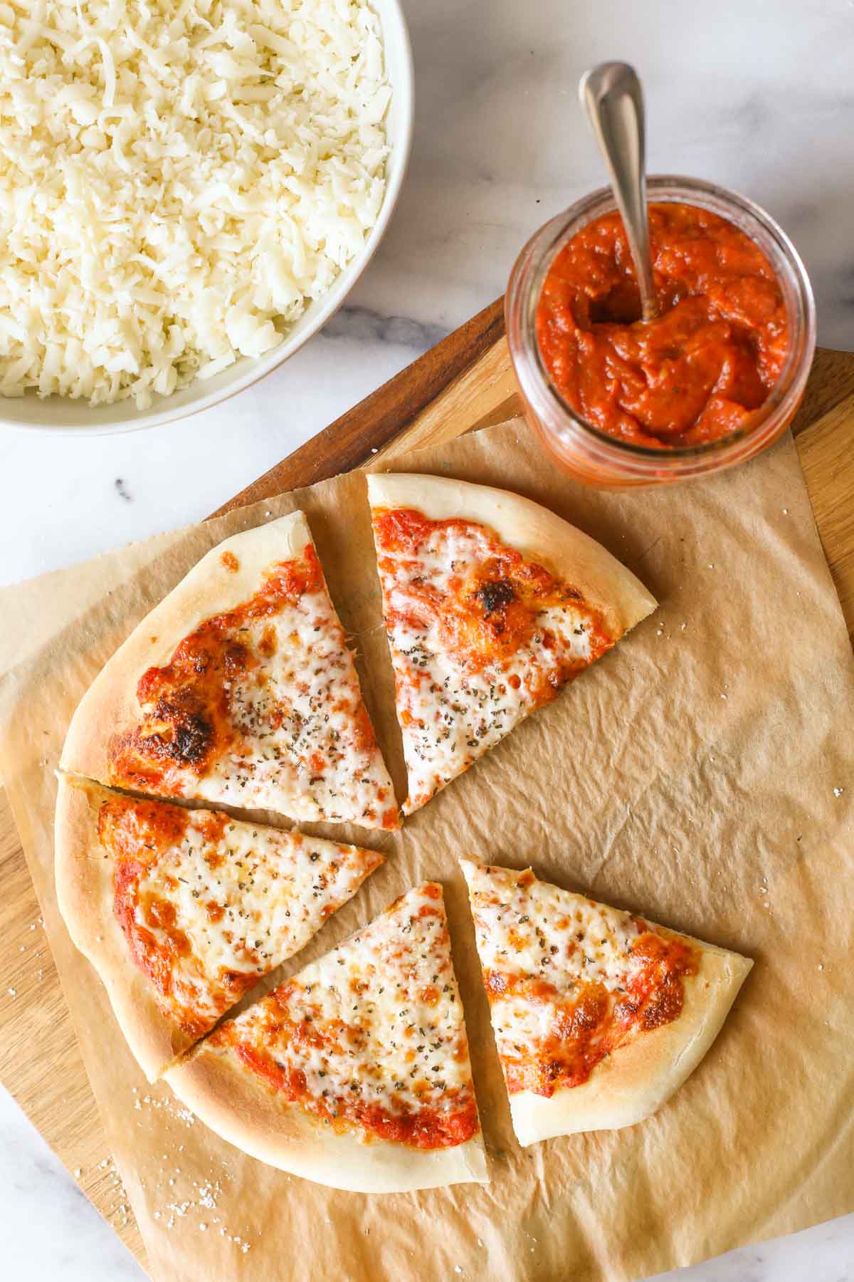 Overhead shot of slices of homemade cheese pizza on parchment paper, sitting on a wood cutting board with a jar of Roasted Tomato Pizza Sauce, and a bowl of shredded mozzarella cheese next to the cutting board. 