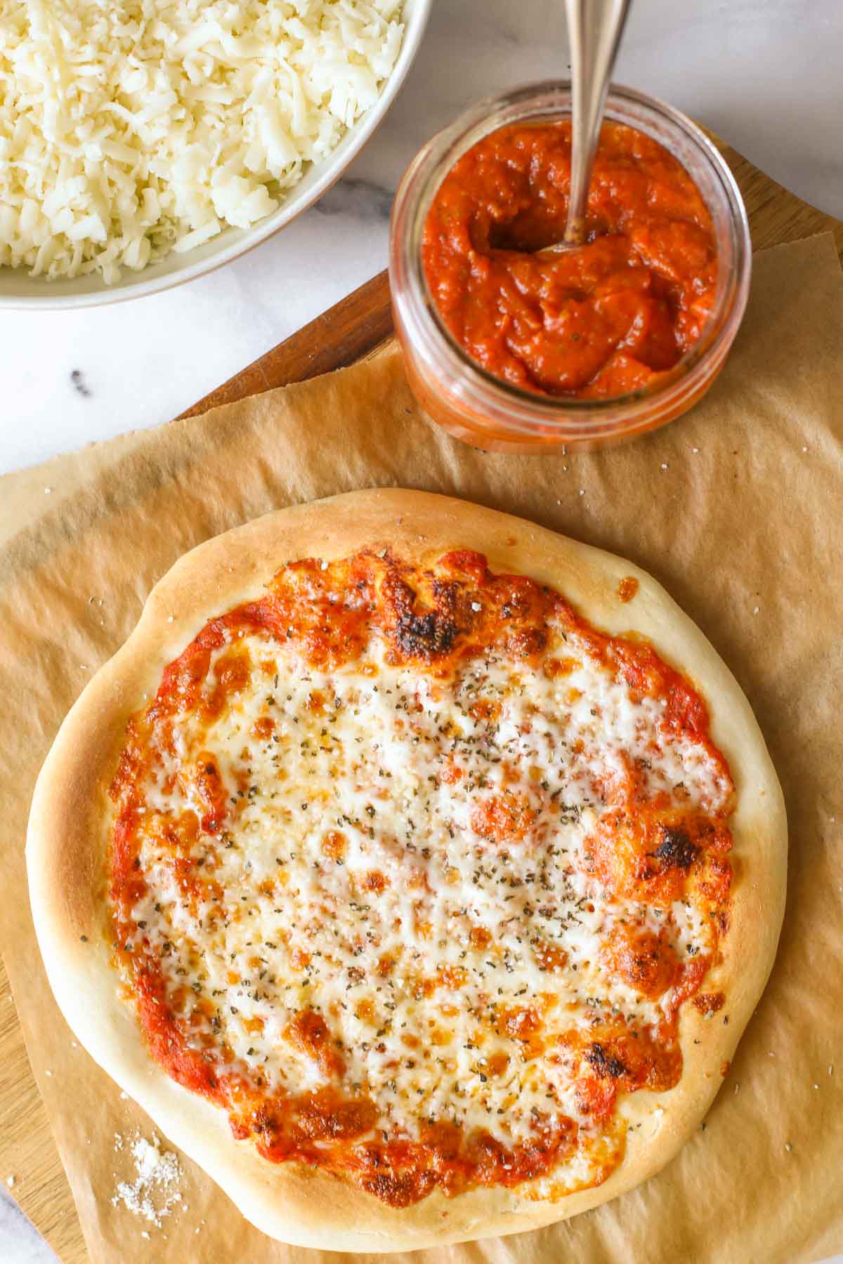 Overhead shot of a homemade cheese pizza on parchment paper, sitting on a wood cutting board with a jar of Roasted Tomato Pizza Sauce, and a bowl of shredded mozzarella cheese next to the cutting board. 