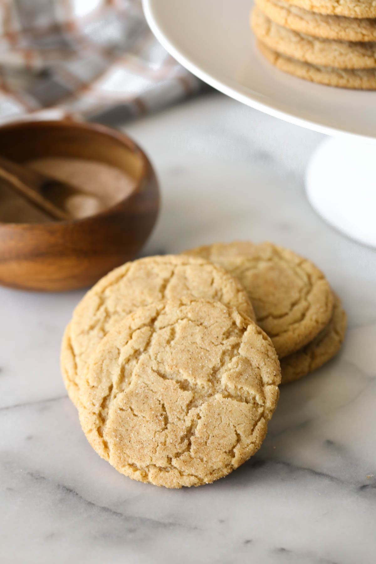A pile of four Brown Butter Snickerdoodles, with a small wood bowl of cinnamon sugar in the background, along with a white cake stand with more cookies stacked on it. 