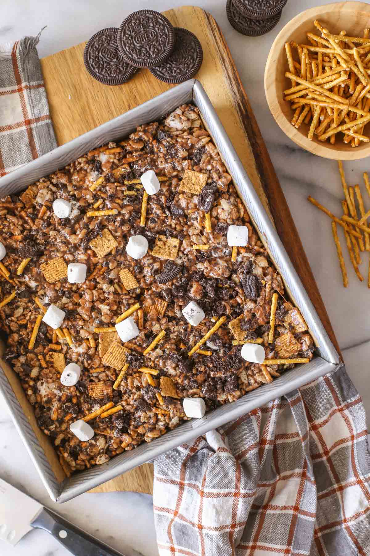 Overhead shot of a square pan of S'more Bars on a wood cutting board, with some whole Oreo cookies and a small wood bowl of pretzel sticks next to it.