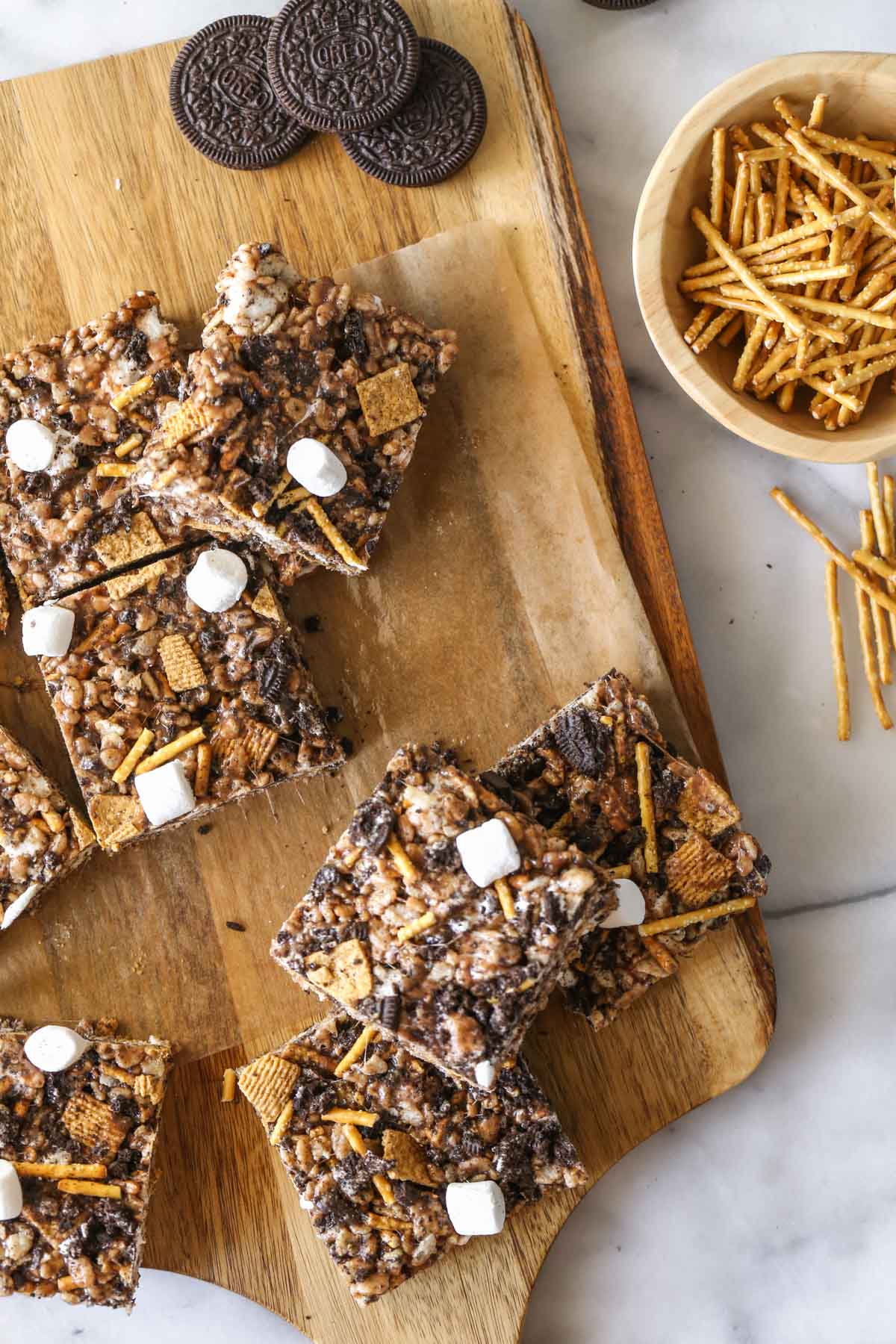 Overhead shot of cut S'more Bars on parchment paper on a wood cutting board, with some whole Oreo cookies and a small wood bowl of pretzel sticks next to it.