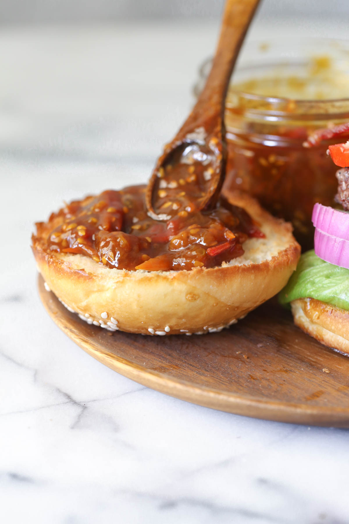 Tomato Jam being spooned onto the top of a hamburger bun with a small wooden spoon, with a small glass jar of Tomato Jam next to the bun.  