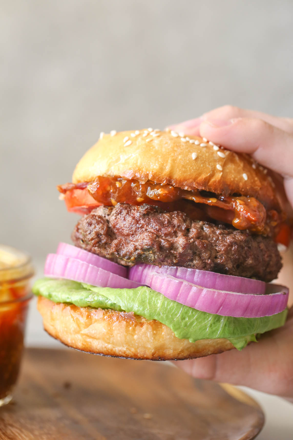 Close up shot of a hand holding a hamburger on a bun with lettuce, red onion, tomato, bacon and Tomato Jam.  