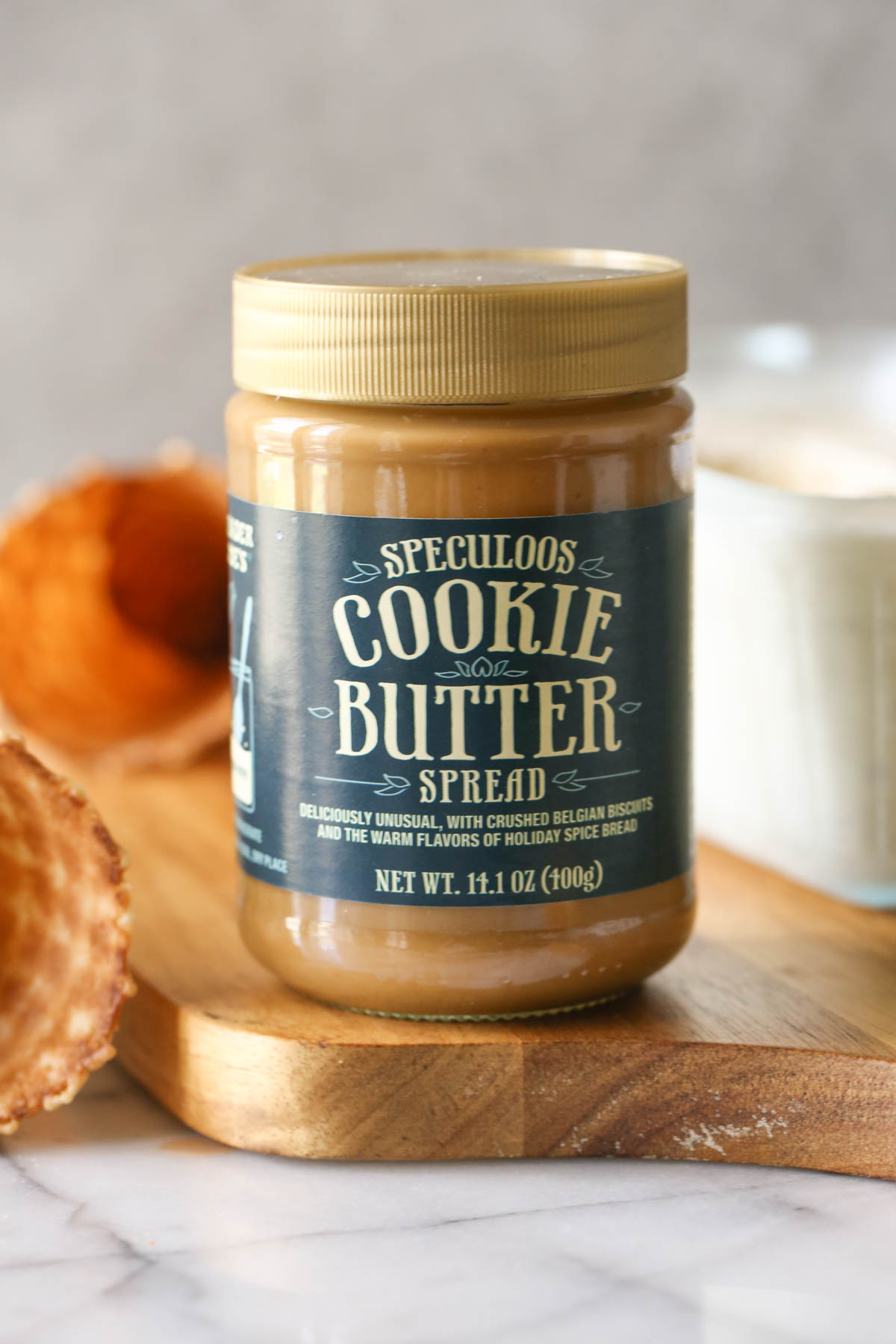 A jar of Speculoos Cooke Butter Spread on a wood cutting board, with waffle cones and a container of Cookie Butter Crisp Ice Cream in the background. 