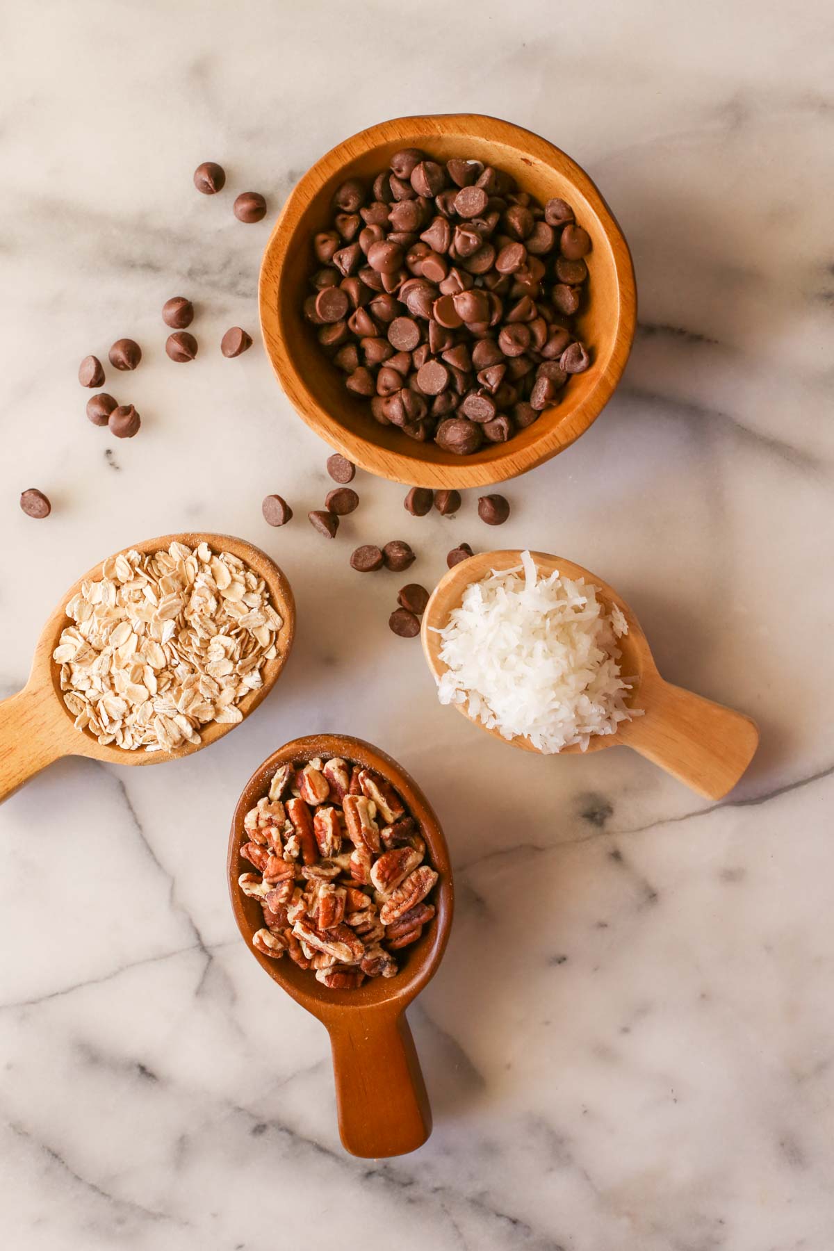 Overhead shot of a wood bowl of chocolate chips, with some of the chocolate chips next to the bowl, a small wood scoop of rolled oats, a small wood scoop of pecans, and a small wood scoop of shredded coconut, all on a marble background. 