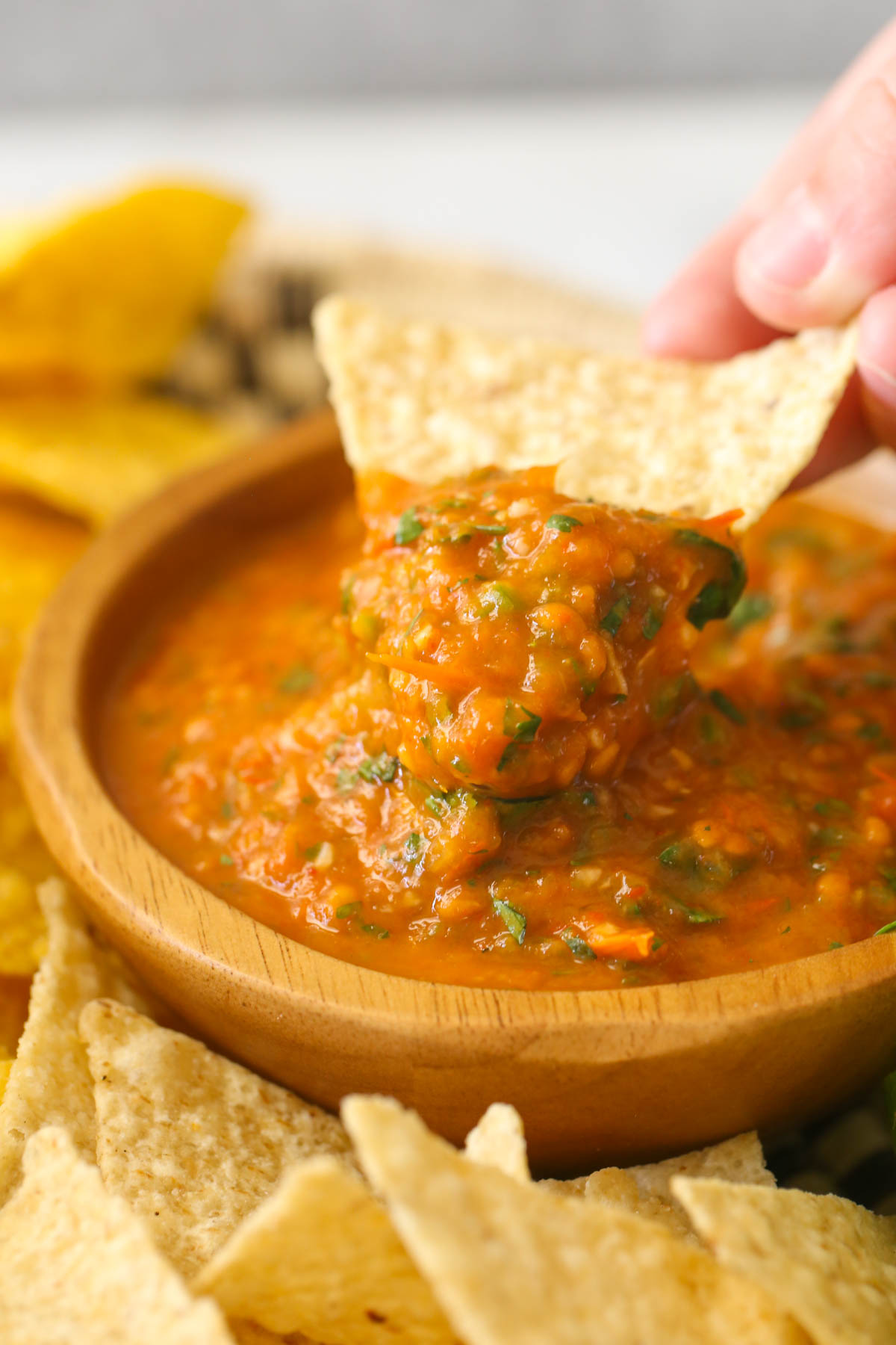 Close up shot of a hand holding a tortilla chip with Roasted Tomato Salsa on it above a wood bowl of Roasted Tomato Salsa with tortilla chips surrounding it. 