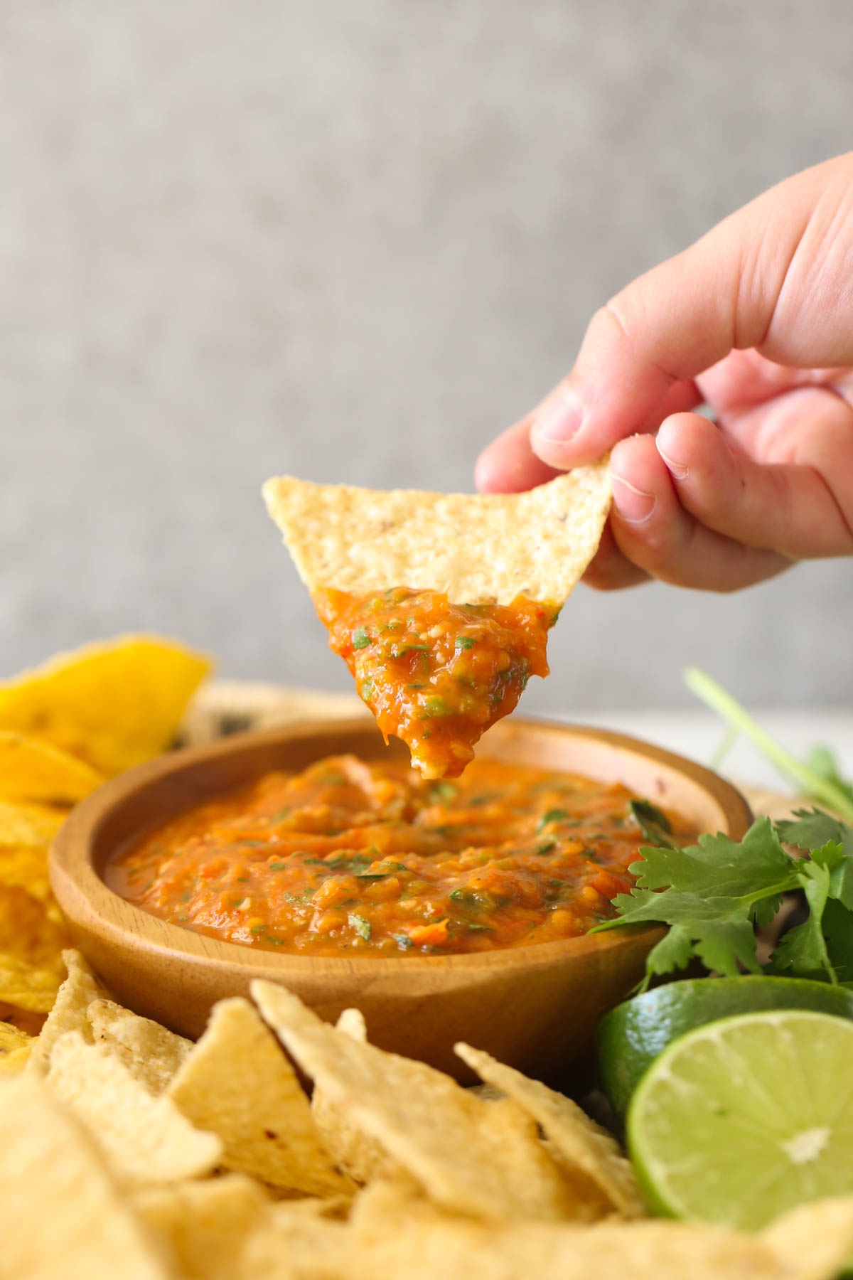 A hand holding a tortilla chip with Roasted Tomato Salsa on it above a small wood bowl of Roasted Tomato Salsa with tortilla chips, two lime halves and fresh cilantro next to the bowl. 