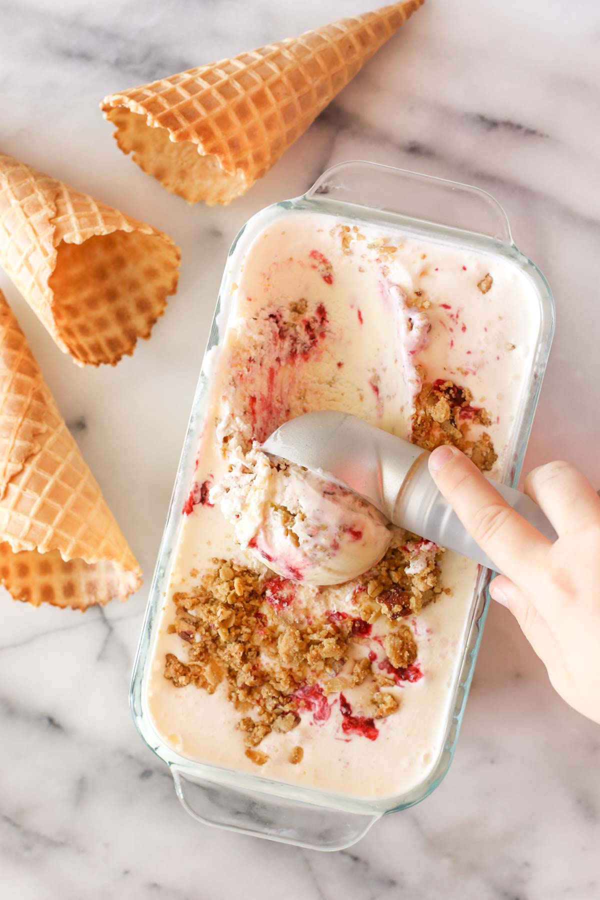 Overhead shot of a hand using an ice cream scoop in a glass container of White Chocolate Raspberry Crisp Ice Cream, with three waffle cones next to the container of ice cream, all on a marble background. 