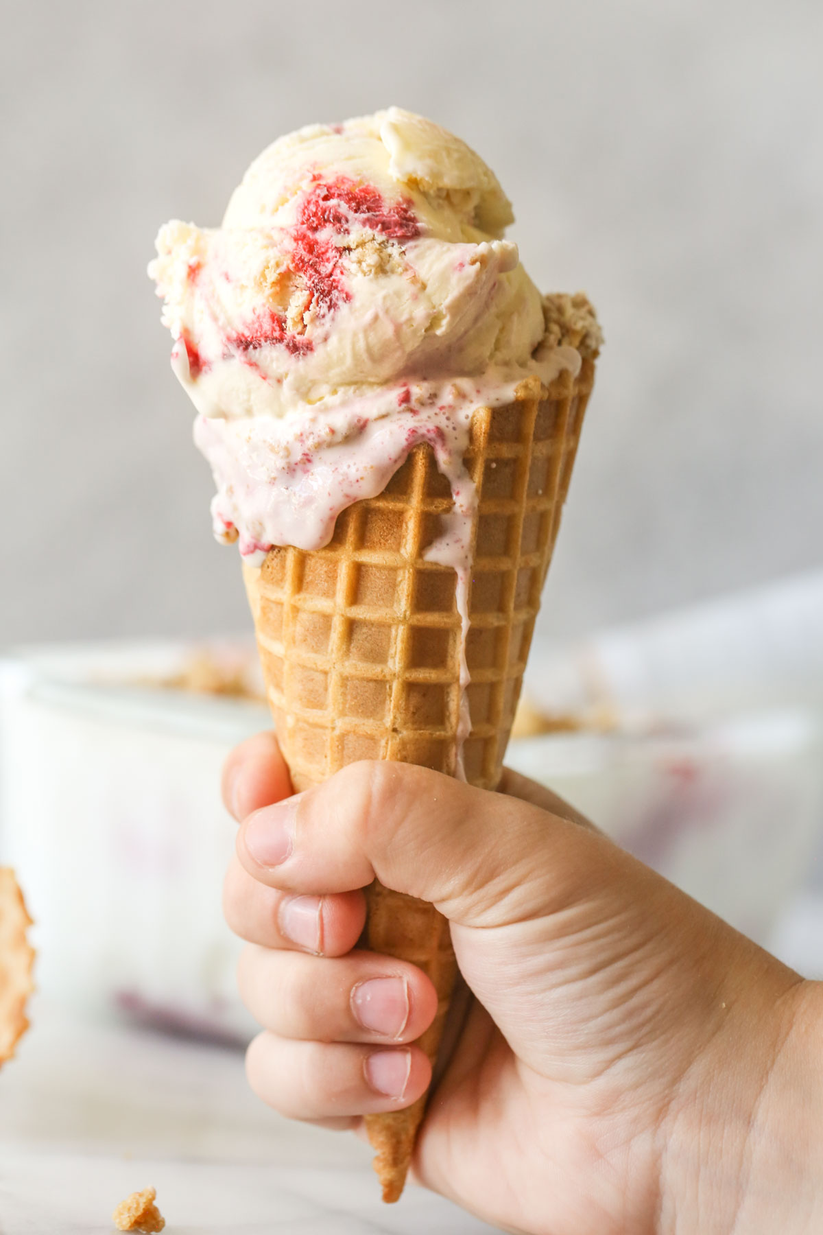 A hand holding a waffle cone with scoops of White Chocolate Raspberry Crisp Ice Cream.  