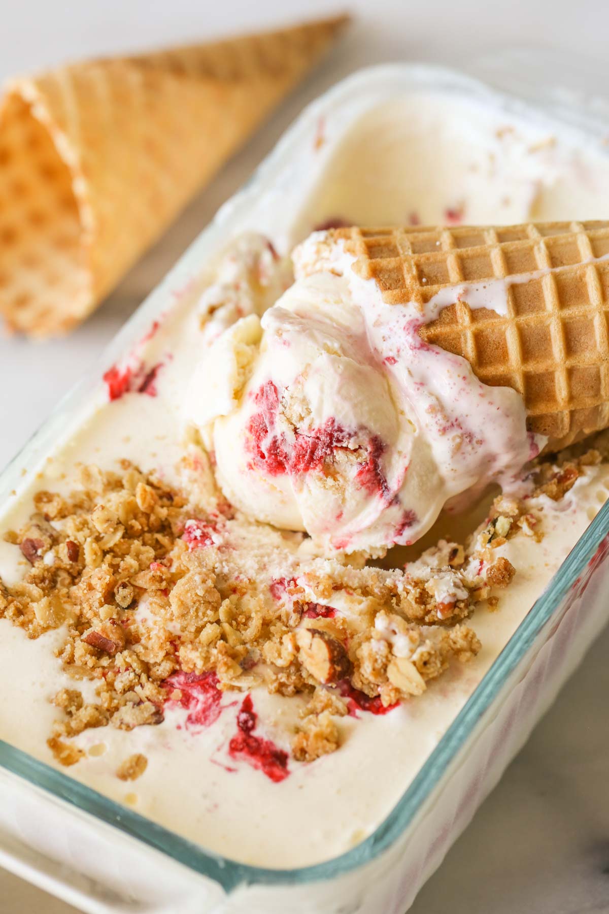 Close up shot of a waffle cone with White Chocolate Raspberry Crisp Ice Cream in it, placed on top of more White Chocolate Raspberry Crisp Ice Cream in a glass container. 