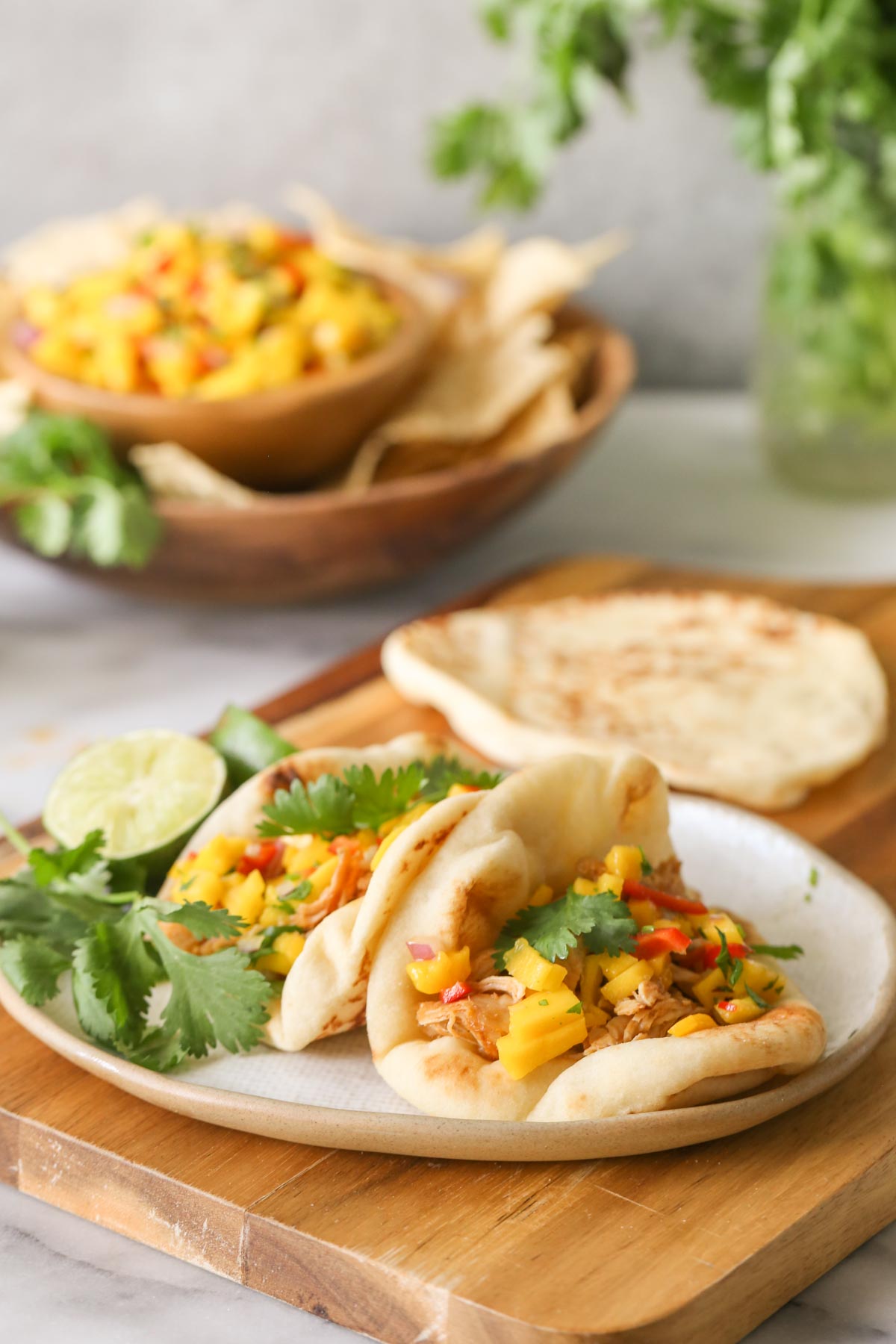 Two pieces of naan bread filled with Buttery Honey Chipotle Chicken and topped with Mango Lime Salsa on a plate with a lime sliced in half and fresh cilantro, sitting on a wood cutting board with another piece of toasted naan bread, and a wood bowl of tortilla chips, fresh cilantro and a bowl of Mango Lime Salsa in the background.  