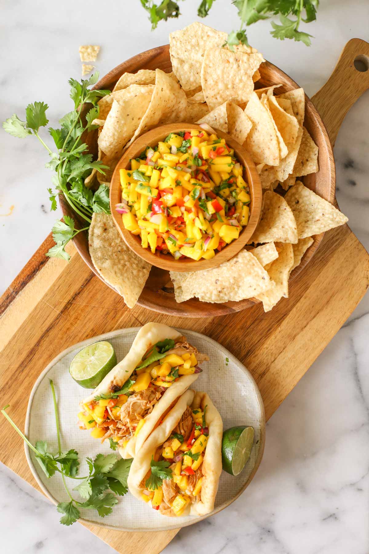 Overhead shot of two pieces of toasted naan bread filled with Buttery Honey Chipotle Chicken and topped with Mango Lime Salsa on a plate with a lime sliced in half and fresh cilantro, sitting on a wood cutting board next to a wood bowl of tortilla chips, fresh cilantro and a bowl of Mango Lime Salsa.  