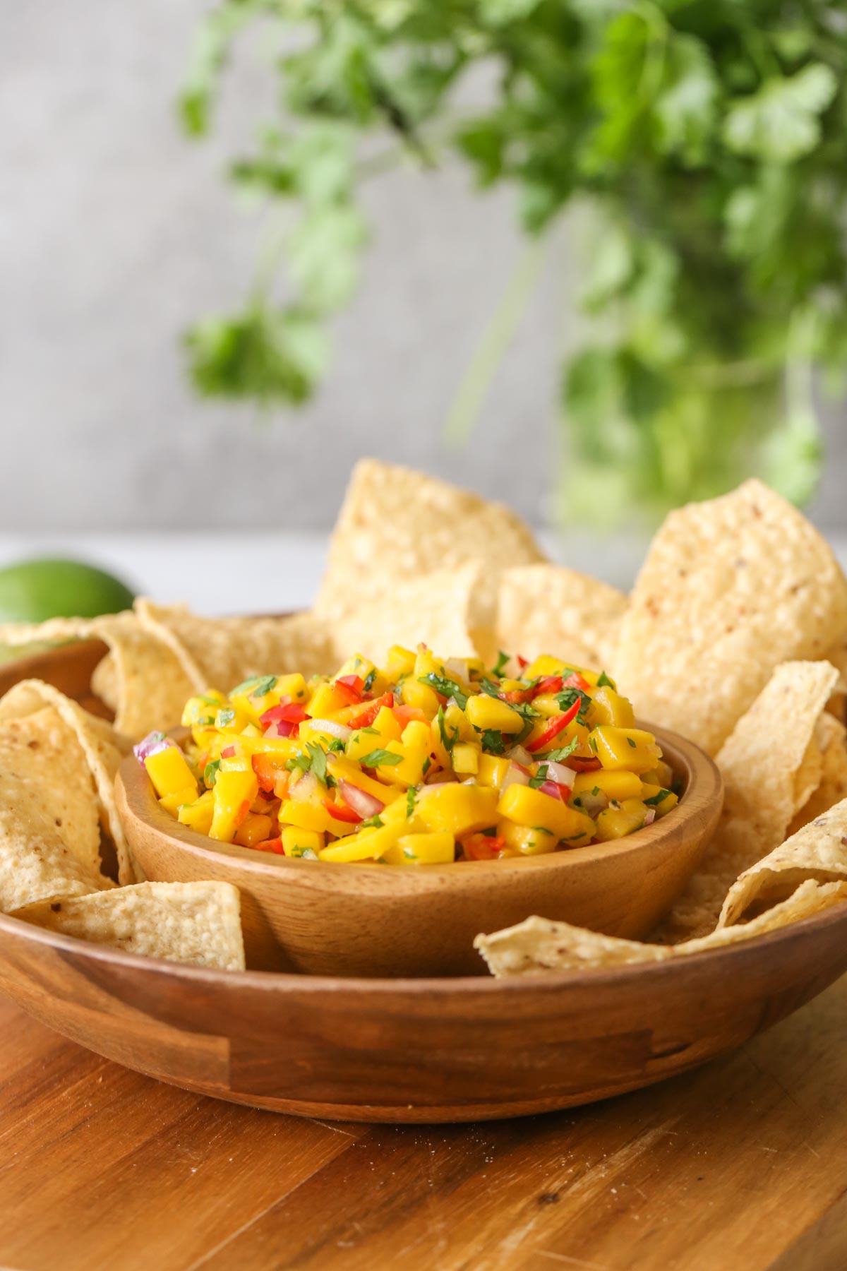 A wood bowl of Mango Lime Salsa sitting inside a larger wood bowl with tortilla chips, all sitting on a wood cutting board.
