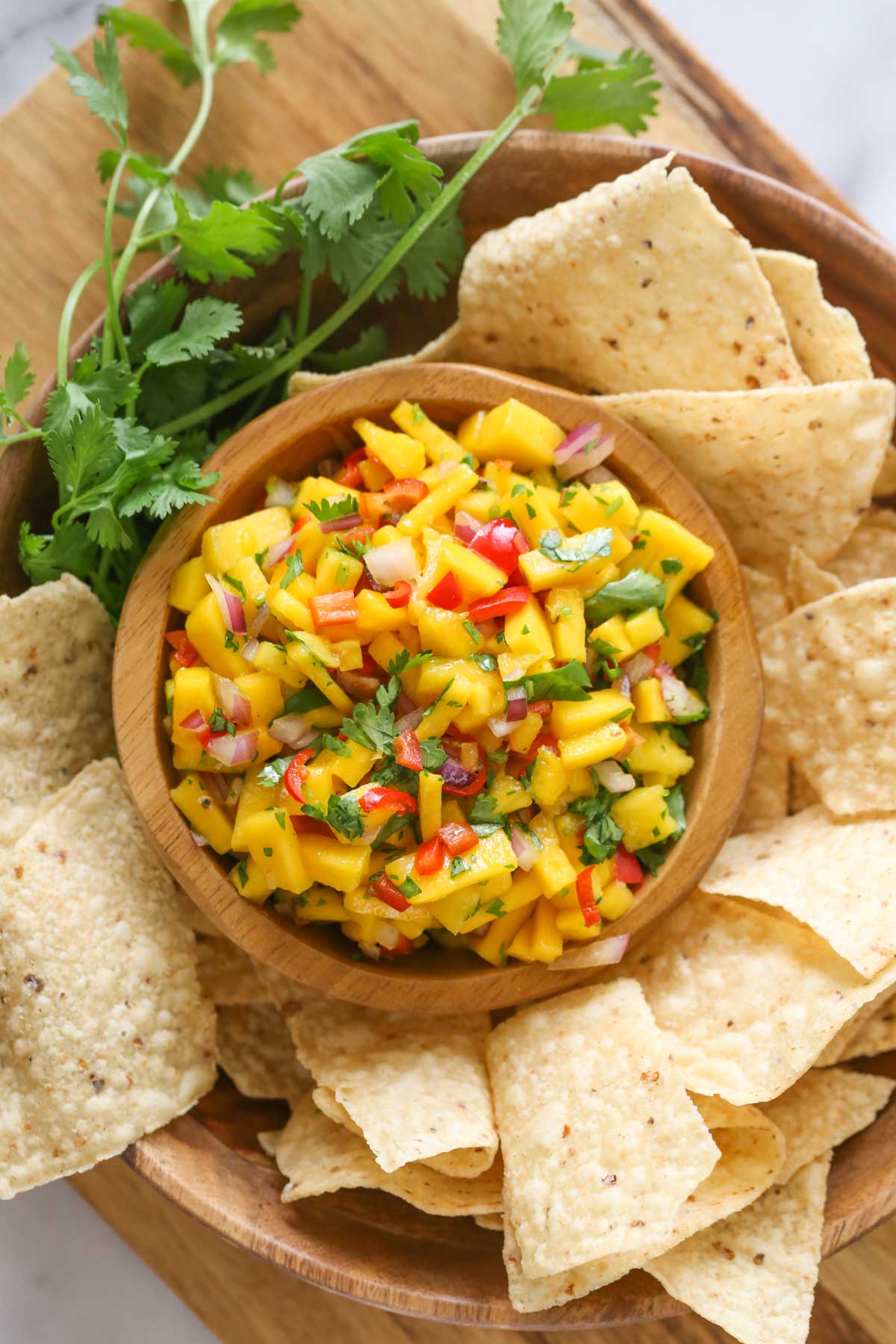 Overhead shot of a wood bowl of Mango Lime Salsa sitting inside a larger wood bowl with tortilla chips and fresh cilantro, all sitting on a wood cutting board.
