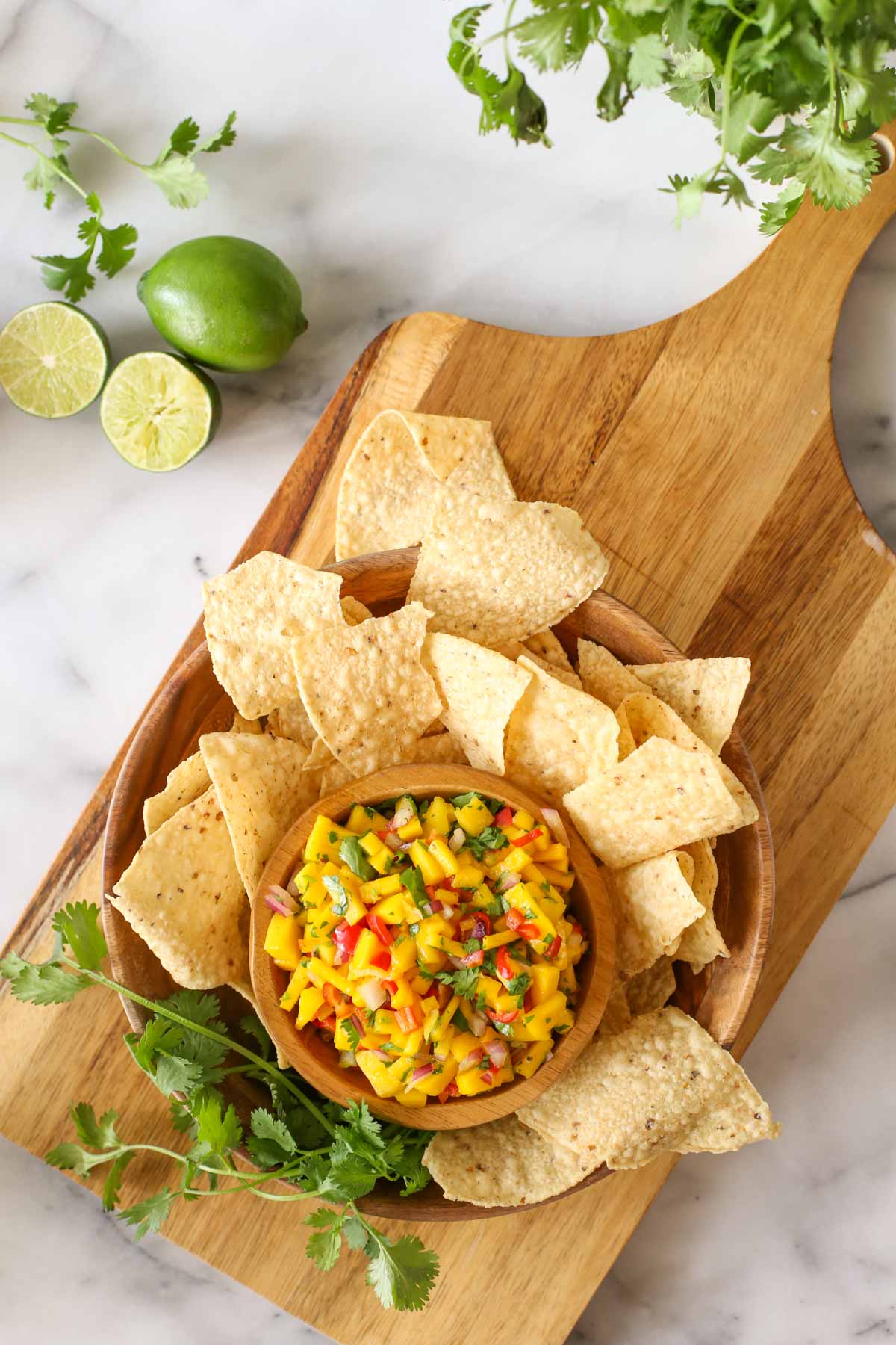 Overhead shot of a wood bowl of Mango Lime Salsa sitting inside a larger wood bowl with tortilla chips and fresh cilantro, all sitting on a wood cutting board with limes and cilantro next to it, all on a marble background. 