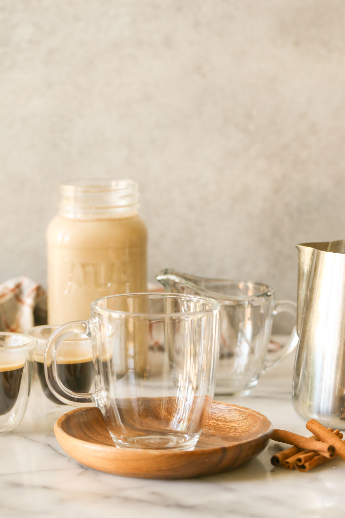 A glass mug on a wood saucer with cinnamon sticks next to it, espresso shots in the background, along with a glass jar of Brown Sugar and Cinnamon Coffee Creamer, a glass pouring mug and a stainless steel pitcher of steamed milk.  