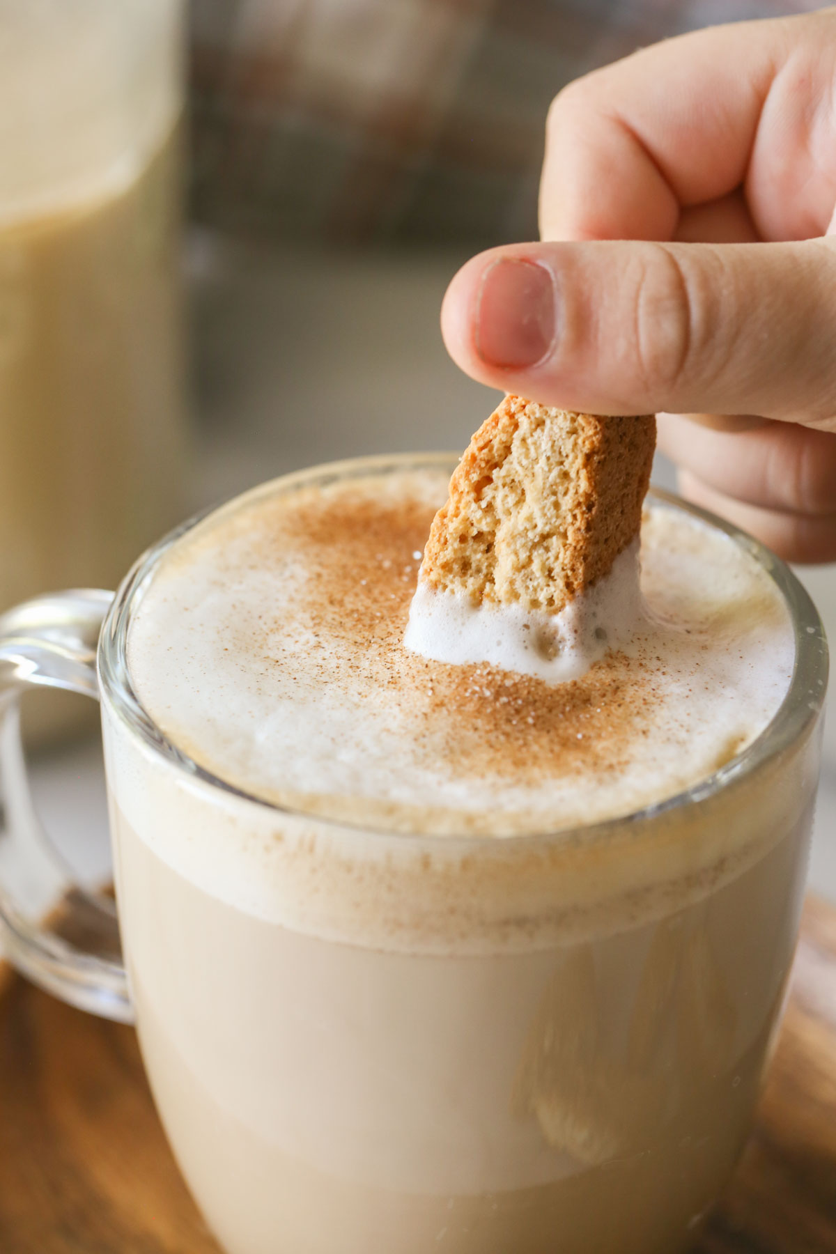 A close up shot of a hand dipping biscotti into a latte in a glass mug, that has Brown Sugar and Cinnamon Coffee Creamer in it and has been dusted with cinnamon and sugar.