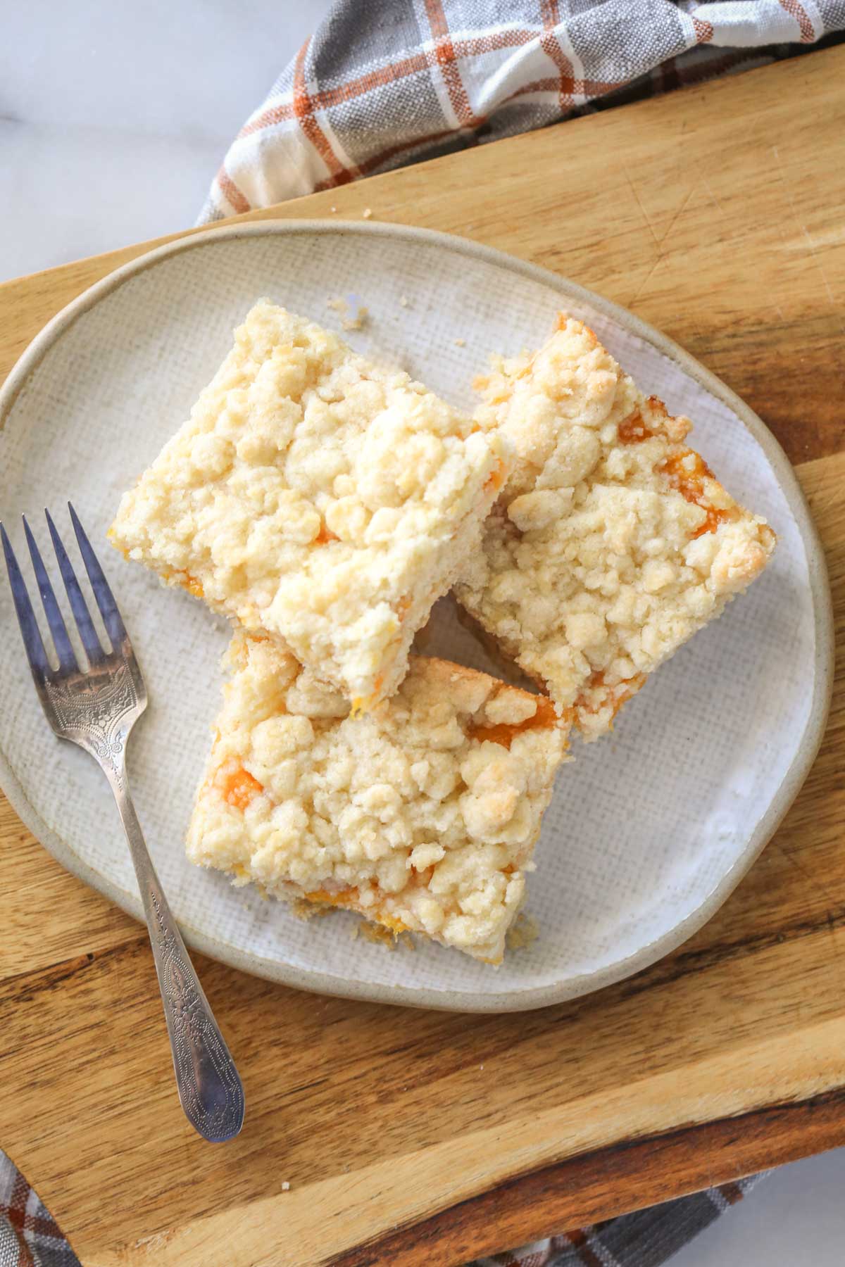 Overhead shot of three Peach Crumb Bar squares on a plate with a fork, sitting on a wood cutting board. 