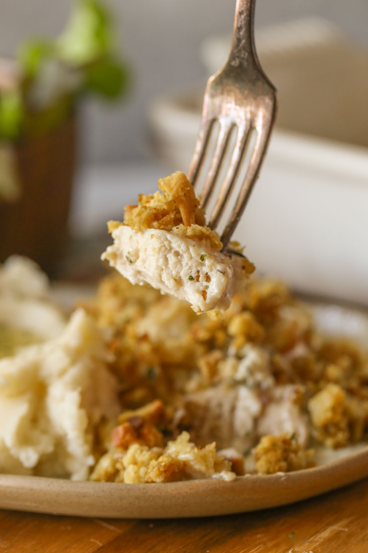 Close up shot of a fork with a bite of Chicken and Stuffing Bake, with a plate of mashed potatoes and Chicken and Stuffing Bake in the background. 