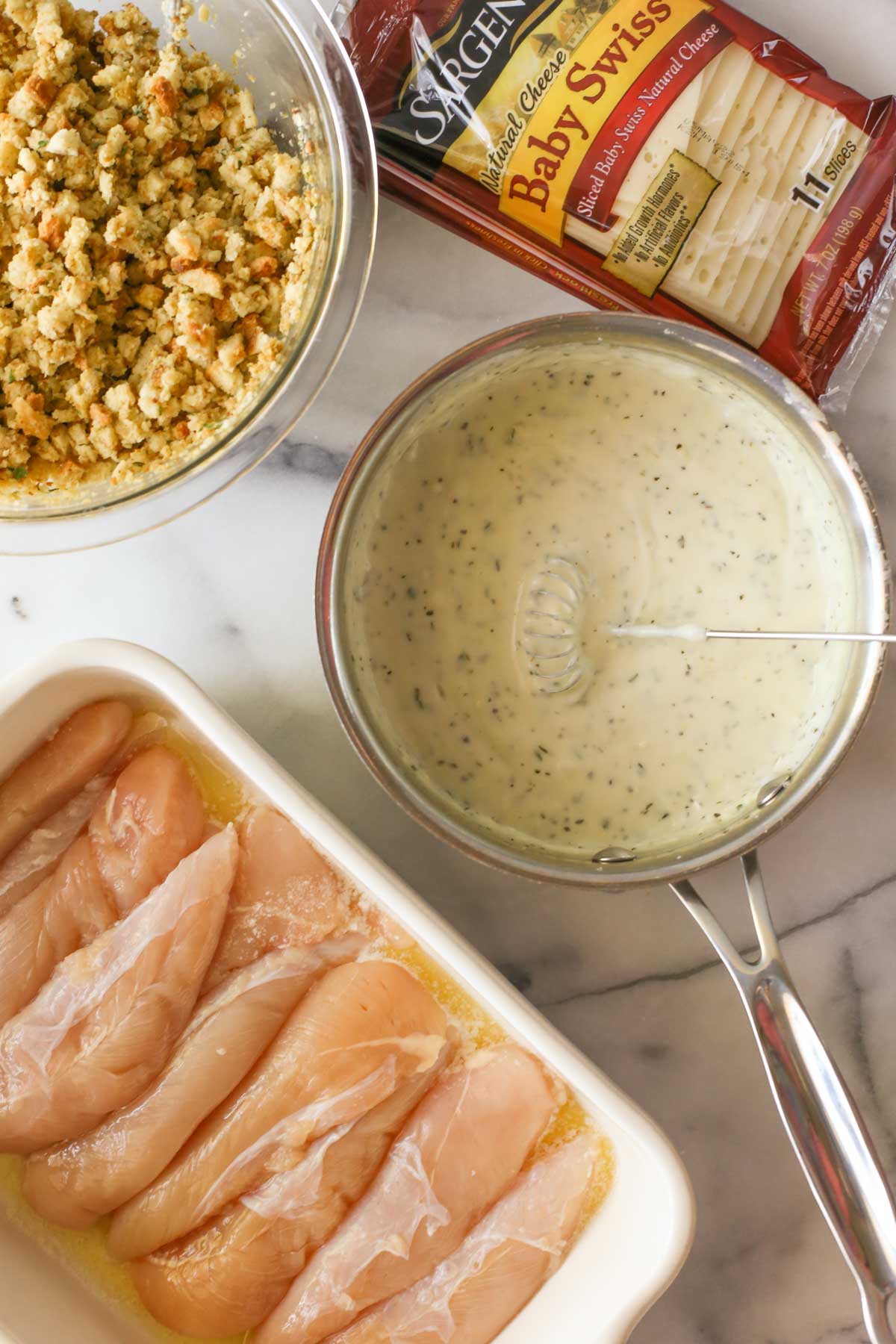 Overhead shot of a sauce pan of Homemade Cream of Chicken Soup, a package of Baby Swiss cheese slices, a mixing bowl of the boxed stuffing, and a baking dish with the melted butter and chicken tenderloins. 