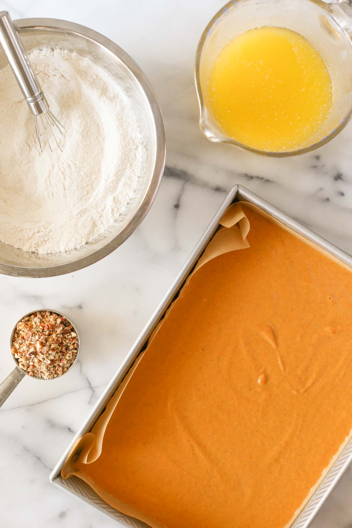 Overhead shot of a parchment paper line baking pan with the pumpkin mixture in it, with a measuring cup of chopped pecans, a mixing bowl of the dried ingredients with a whisk in it, and a measuring cup of melted better all next to the baking pan. 