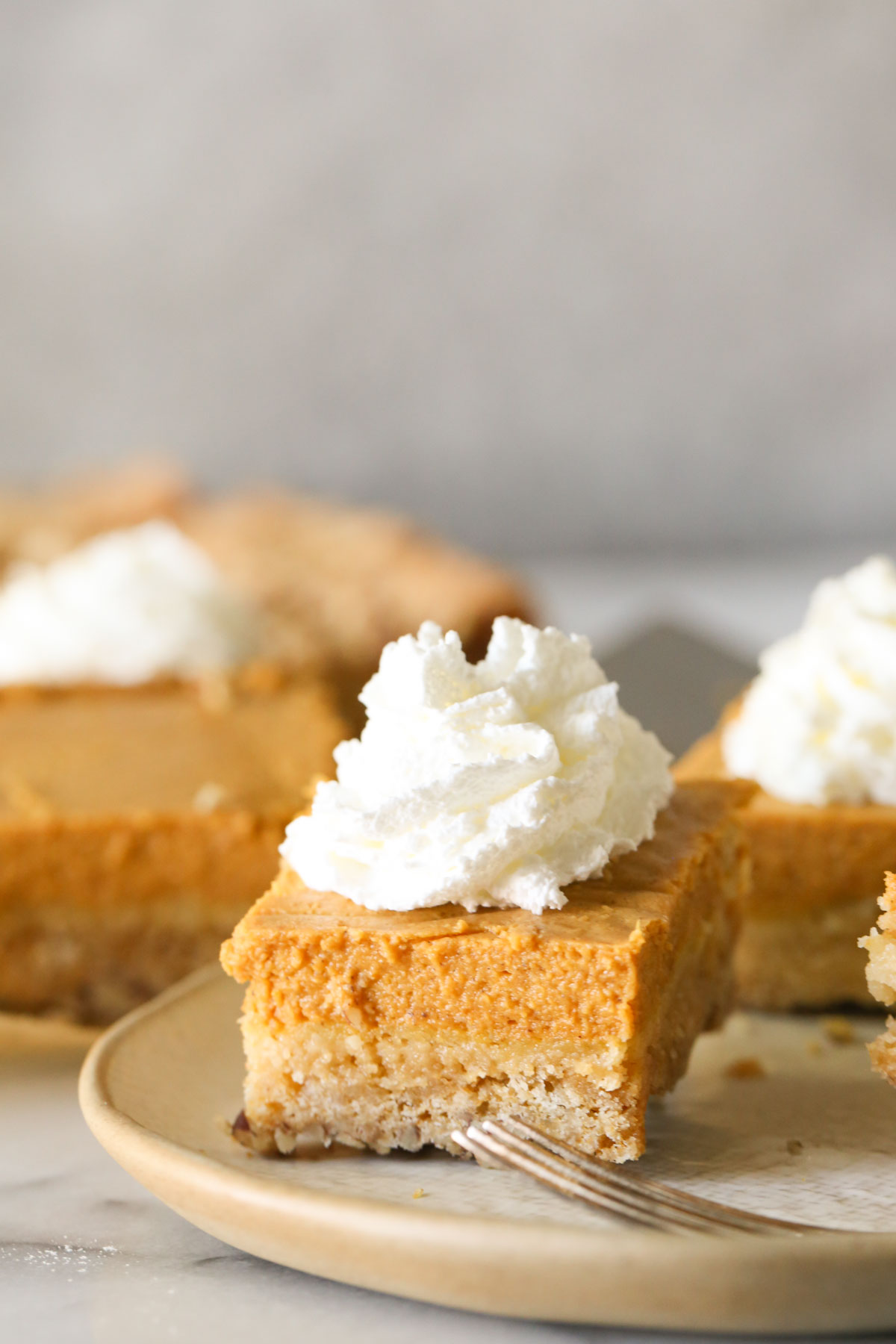 A Pumpkin Pie Bar served upside down on a plate with whipped cream on top, with more Pumpkin Pie Bars in the background. 