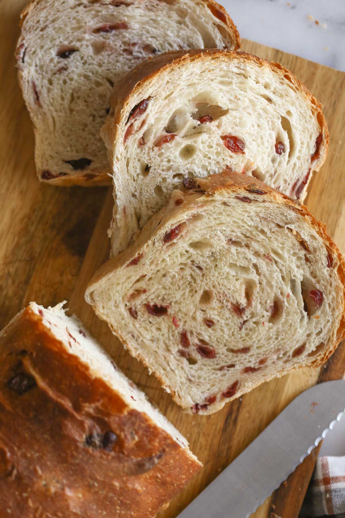 Overhead shot of a loaf of Sourdough Cranberry Bread on a wood cutting board with a serrated knife and three slices of bread that have been cut from the loaf. 