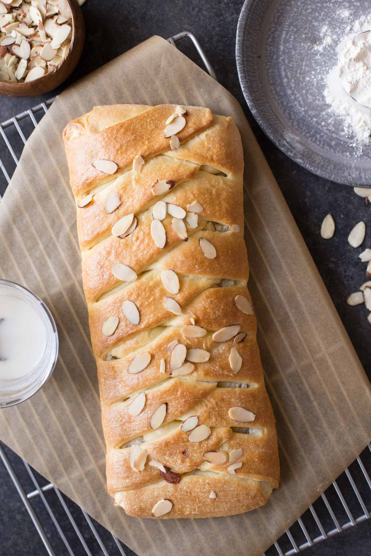 Overhead shot of the Almond Braid on parchment paper on a cooling rack with a small glass bowl of the almond glaze next to it, along with a small wood bowl of sliced almonds and a metal tray with flour next to the cooling rack. 