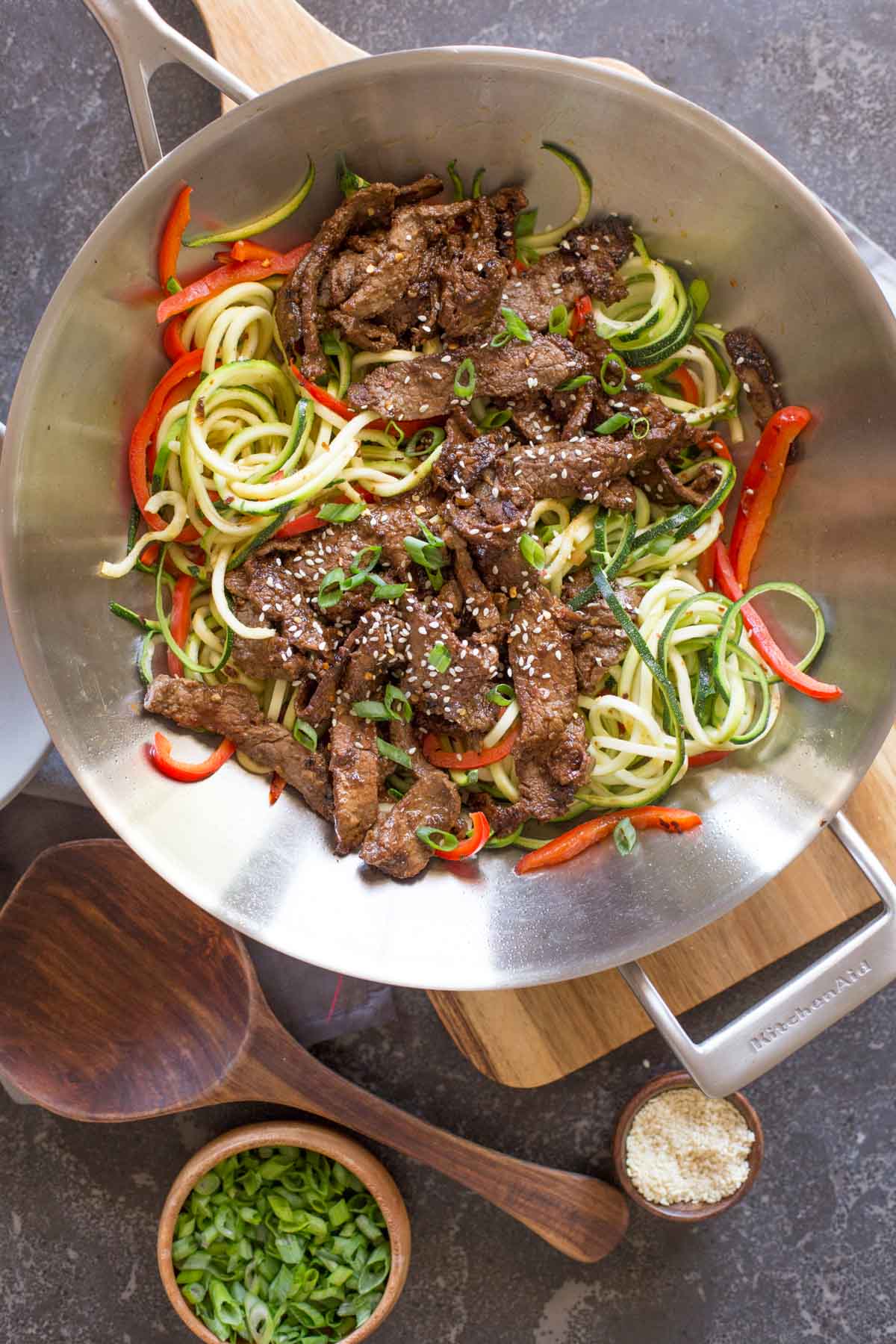 Overhead shot of a large wok with Spicy Korean Beef and Zoodles in it, sitting on a wood cutting board, with a small wood bowl of sesame seeds, a small wood bowl of green onions and a wooden serving spoon next the the cutting board.   