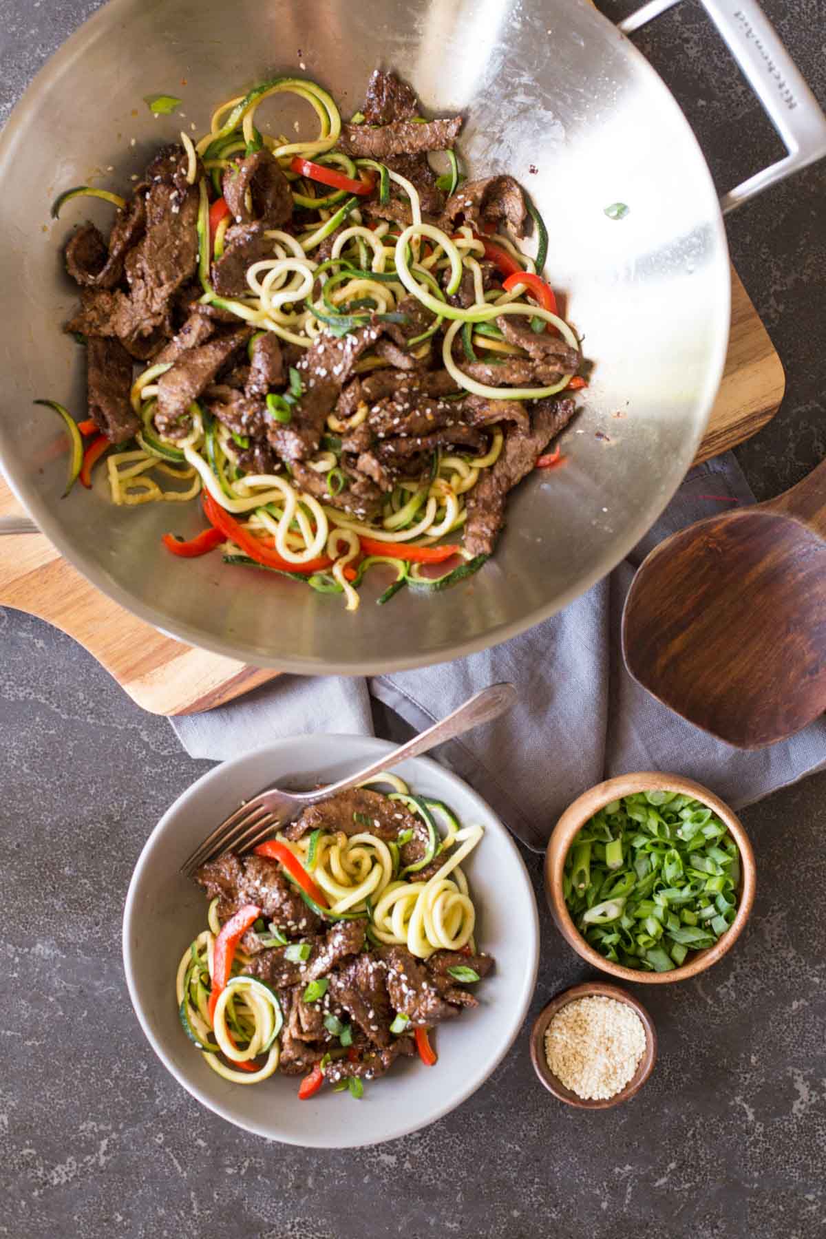 Overhead shot of a large wok with Spicy Korean Beef and Zoodles in it, sitting on a wood cutting board, with a bowl of Spicy Korean Beef and Zoodles next to the wok, along with a small wood bowl of sesame seeds and a small wood bowl of green onions.  