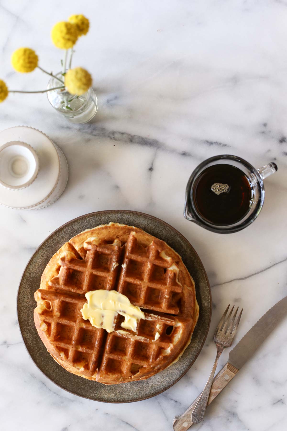 Overhead shot of Overnight Sourdough Waffles on a plate with butter on top, with a mini glass pitcher of syrup, a butter dish, a vase with flowers and silverware all next to the plate. 