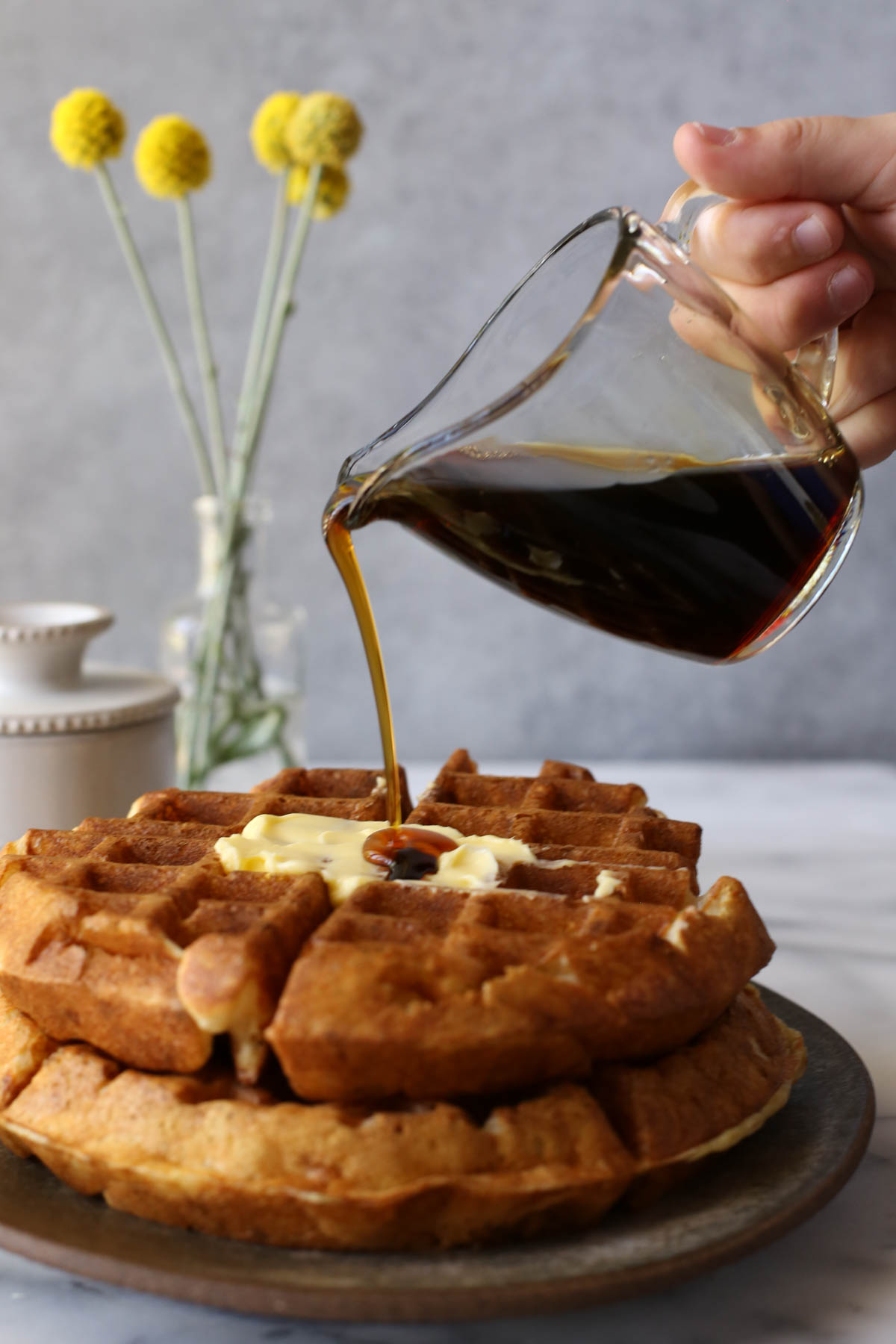 Two Overnight Sourdough Waffles stacked on a plate, with a hand pouring syrup over the waffles from a mini glass pitcher, with a butter dish and flowers in a vase in the background. 
