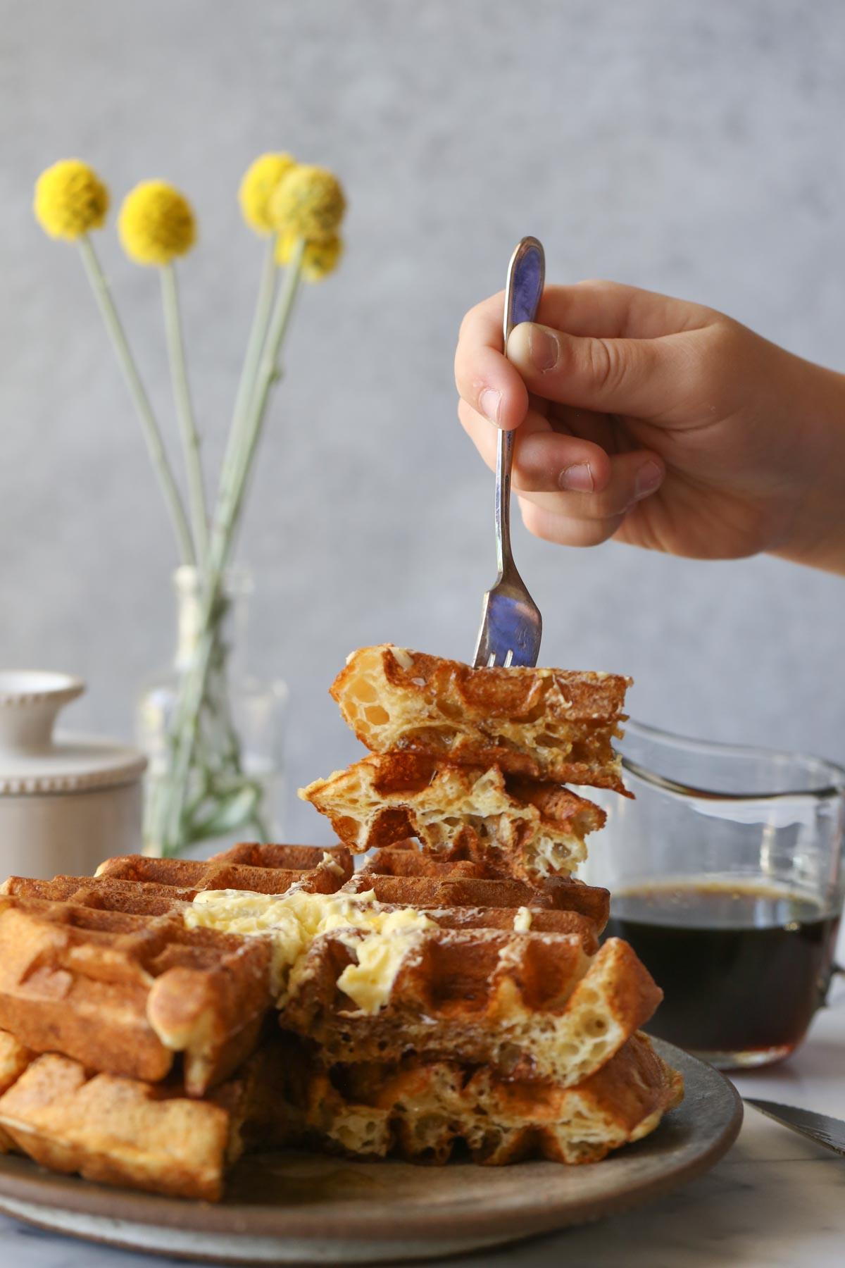 Two Overnight Sourdough Waffles stacked on a plate with a hand holding a fork poked into the waffles, with a mini glass pitcher of syrup, a butter dish and a vase with flowers in the background. 