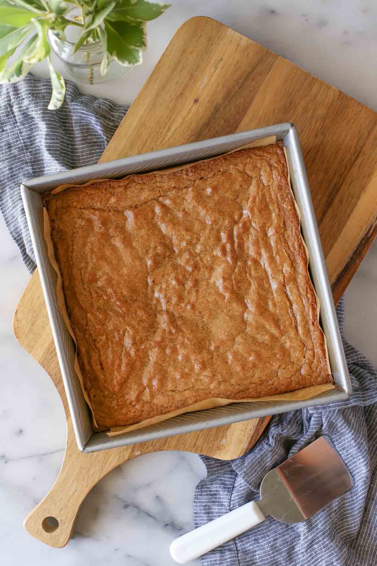 Overhead shot of a square, parchment paper lined baking pan of Best Ever Blondies, sitting on a wood cutting board, with a serving spatula next to it. 