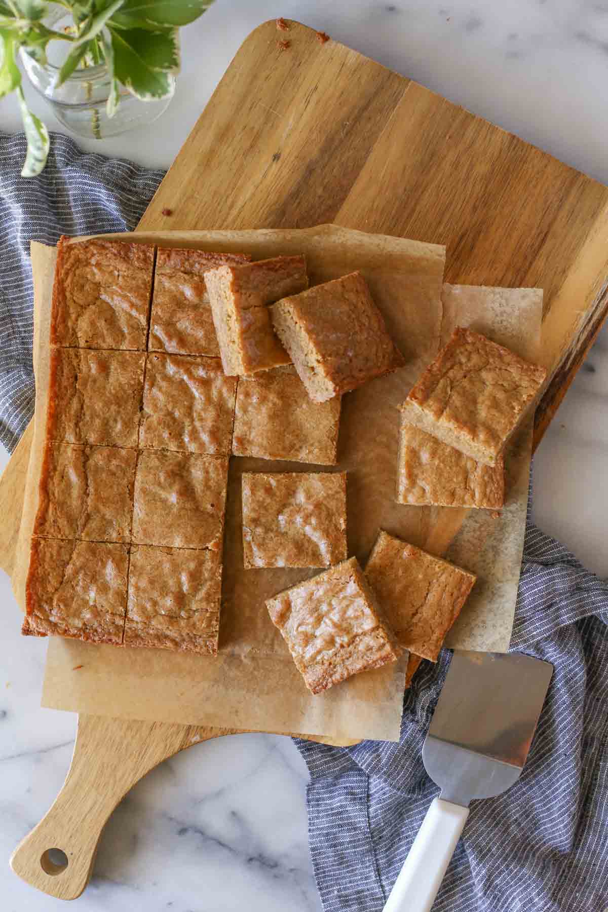 Overhead shot of Best Ever Blondies cut into square bars on parchment paper on top of a wood cutting board, with a serving spatula next to the board. 