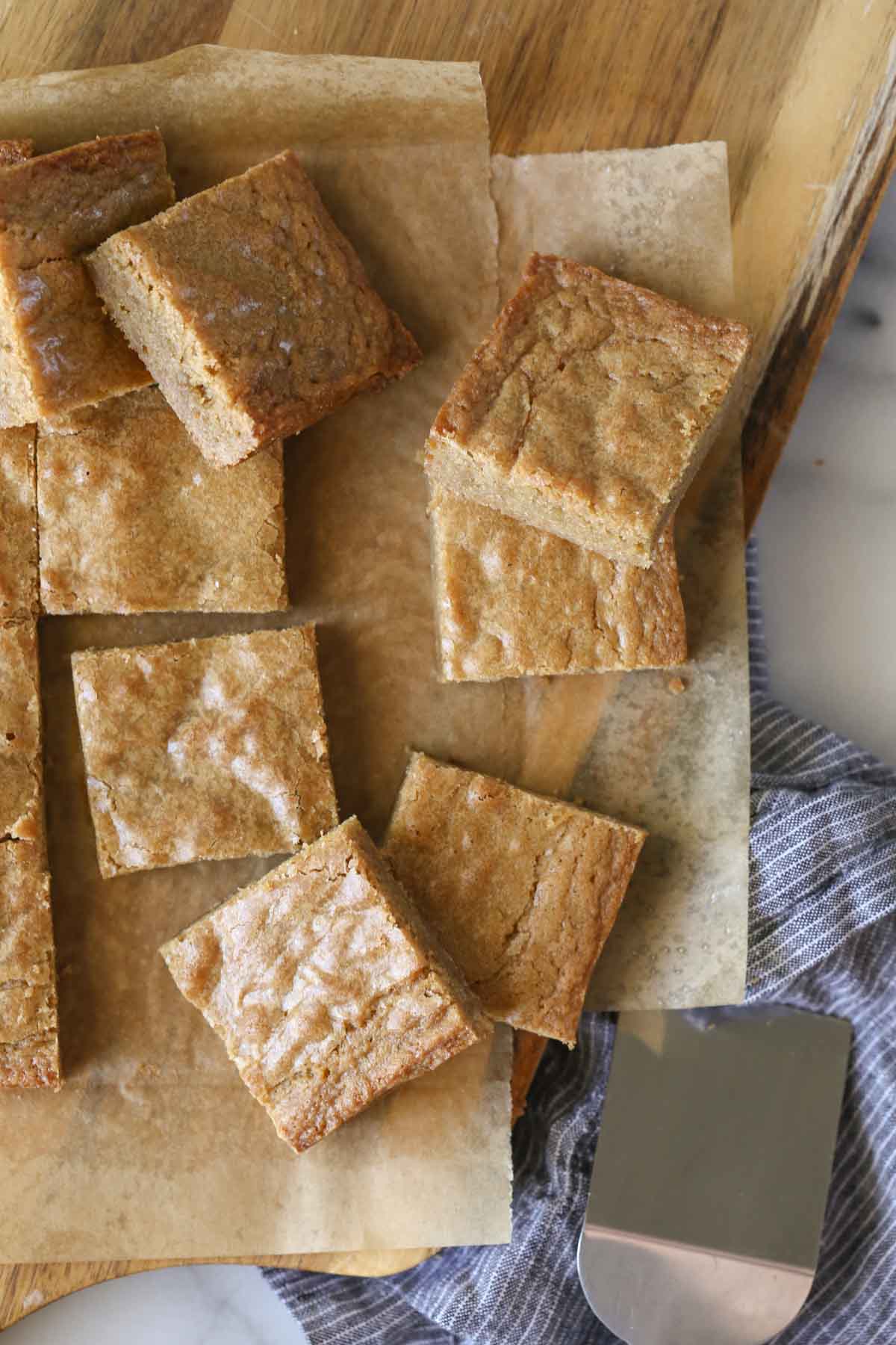 Overhead shot of Best Ever Blondies cut into square bars on parchment paper on top of a wood cutting board, with a serving spatula next to the board. 