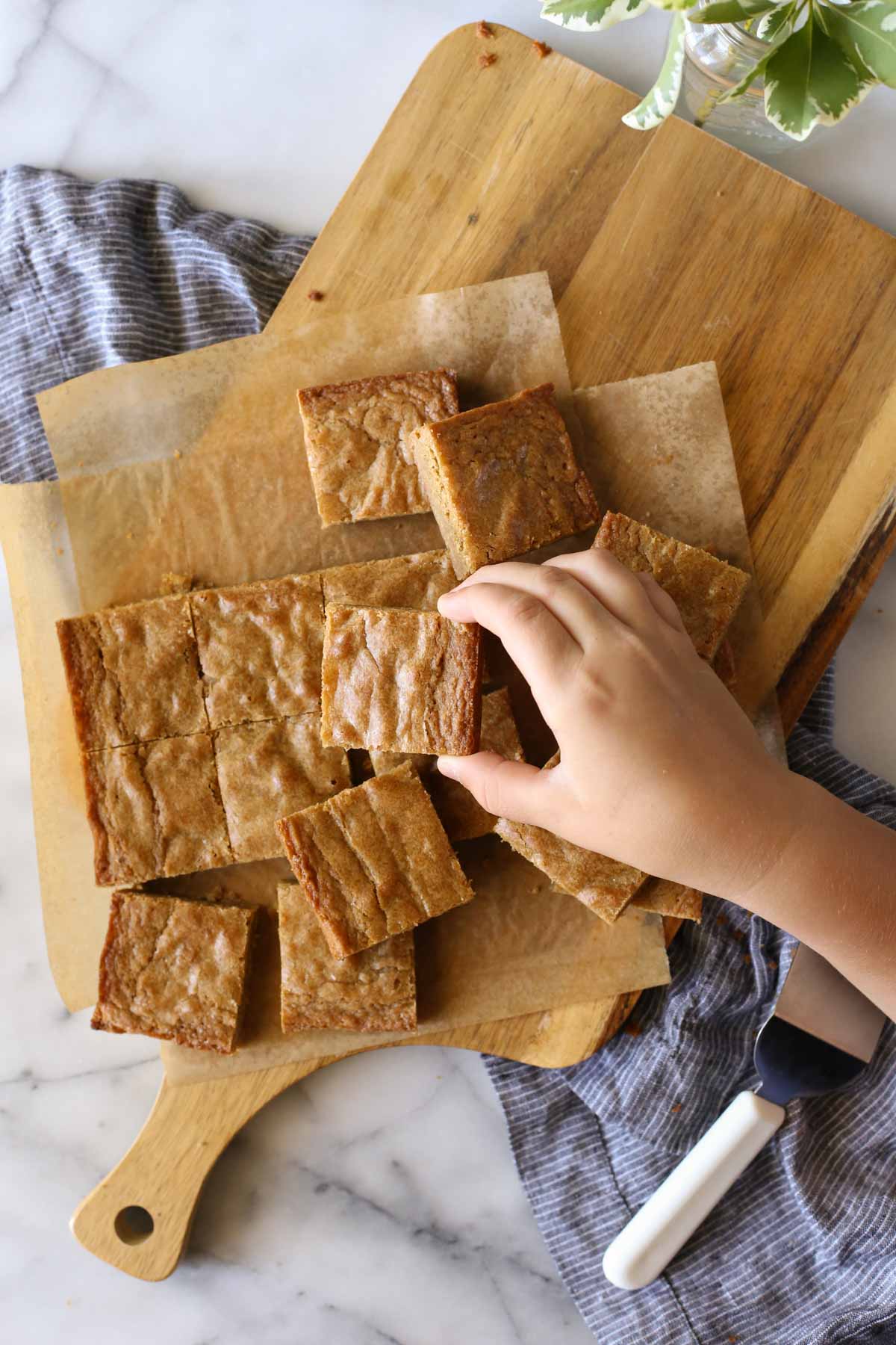 Overhead shot of Best Ever Blondies cut into square bars on parchment paper on top of a wood cutting board, with a hand picking up one of the blondies.   