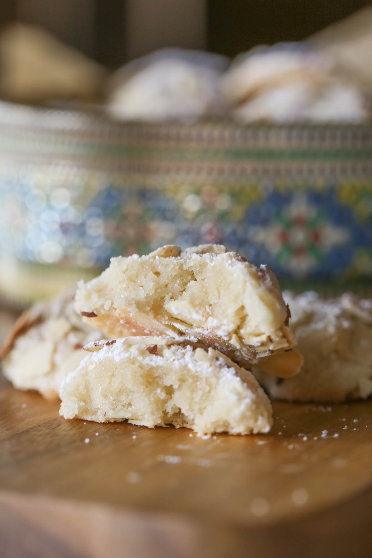 Close up shot of an Almond Paste Cookie broken in half, showing the inside texture, with more cookies in the background along with a cookie tin. 