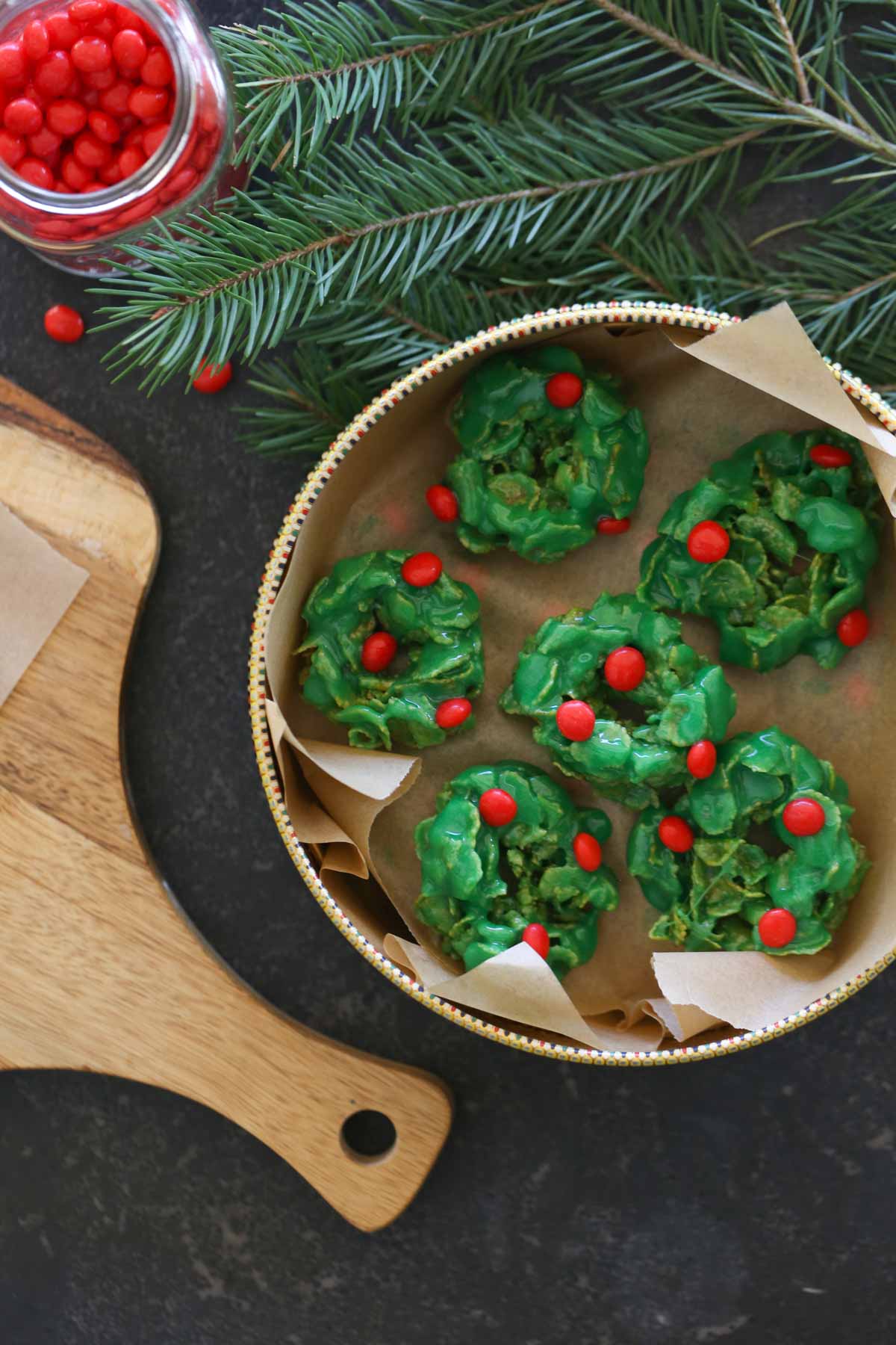 Overhead shot of Holly Cookies in a parchment paper lined cookie tin, sitting next to a wood cutting board and a small glass jar of cinnamon candies. 