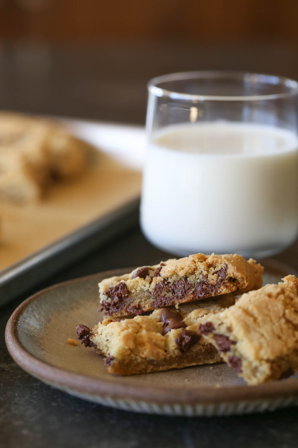 Close up shot of Chocolate Chip Cookie Sticks on a plate, with a glass of milk in the background along with the baking pan of more Chocolate Chip Cookie Sticks. 