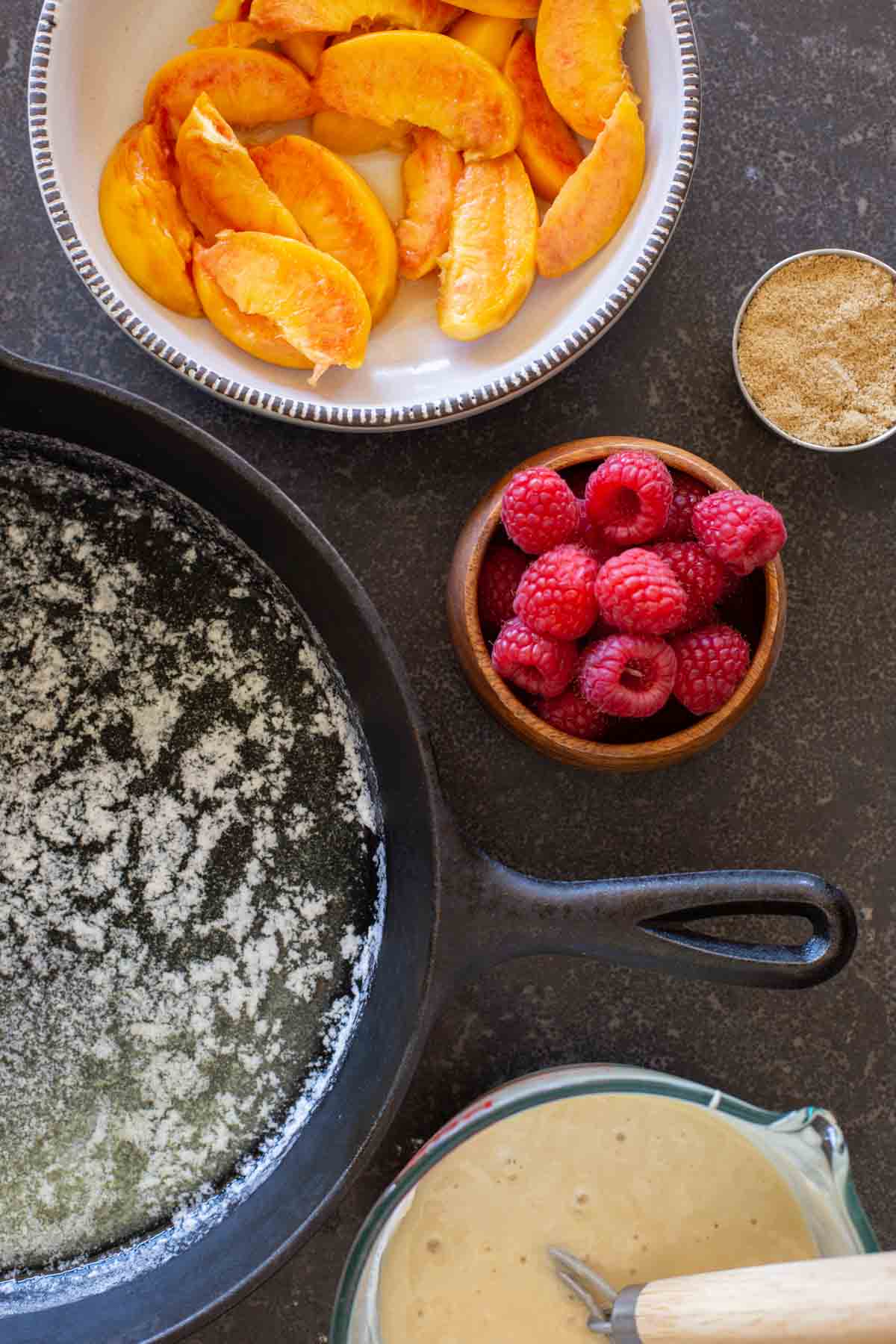 Overhead shot of the ingredients for Sourdough Peach Cobbler - melted butter in a cast iron skillet, a bowl of sliced peaches, a small bowl of raspberries, a cup of brown sugar, and a mixing bowl of the cobbler batter. 