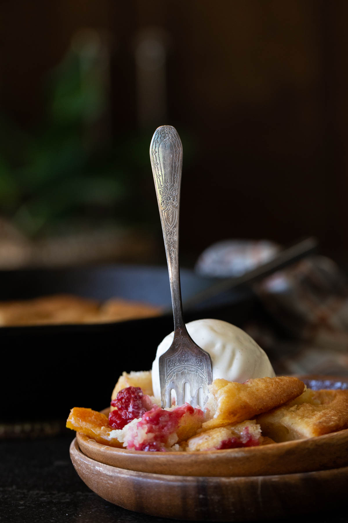 A fork stuck into a serving of Sourdough Peach Cobbler topped with vanilla ice cream in a wood bowl. 
